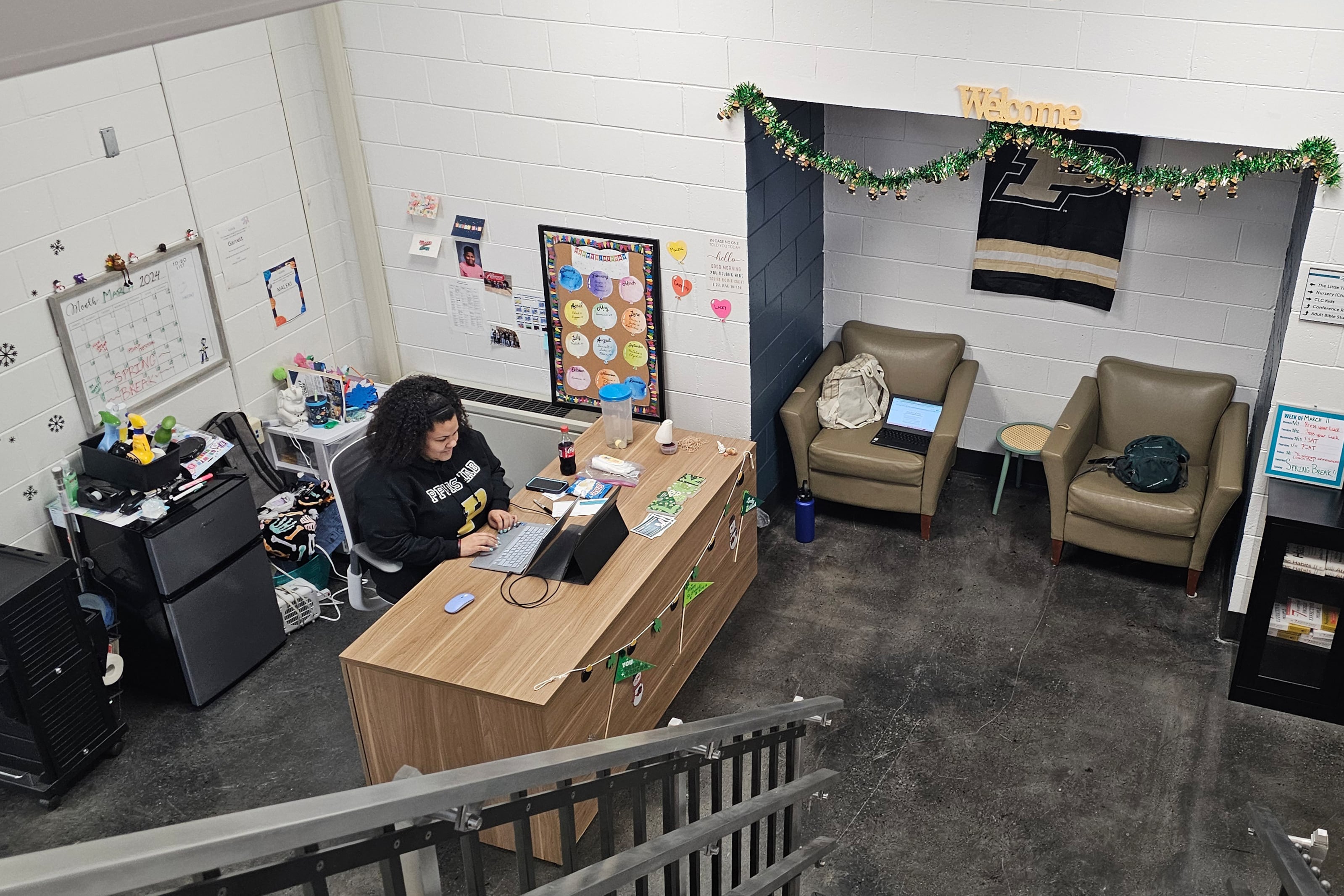 A view from above of an adult with dark hair and wearing a dark sweater sits at a wooden desk typing at a laptop in an office environment.