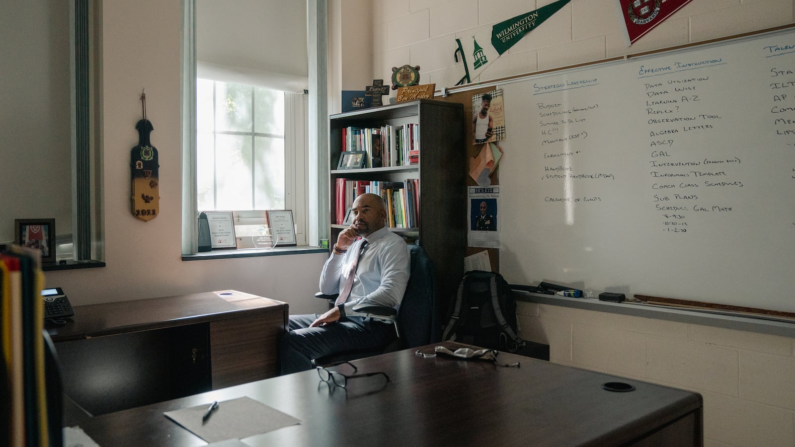A school principal sits at the desk in his office next to a window. 