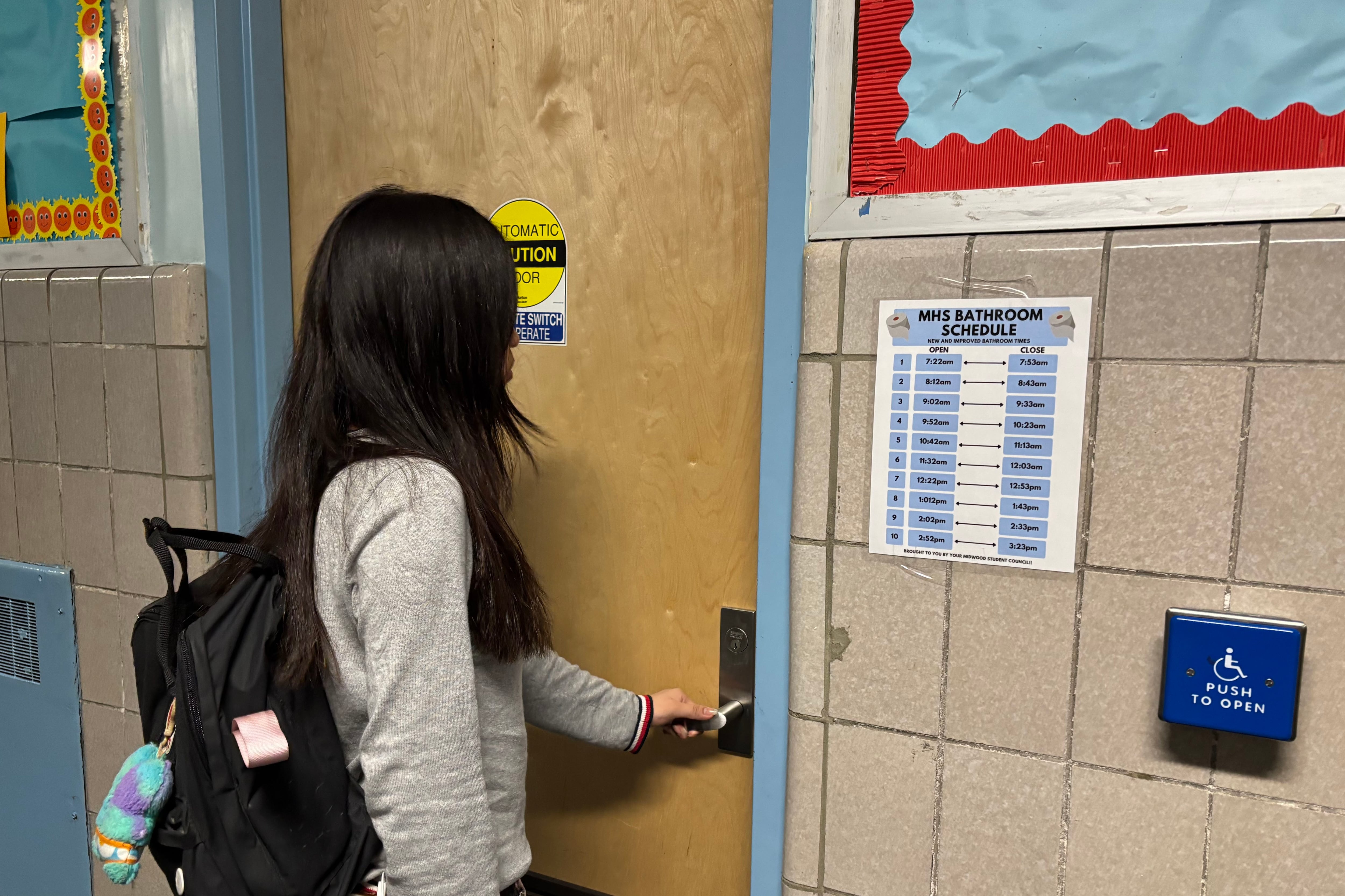 A high school student with long hair and wearing a backpack reaches to open a bathroom door in a school.