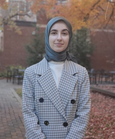 A woman in a green hijab and coat stands smiling for a photo with a red brick building and trees in the background.