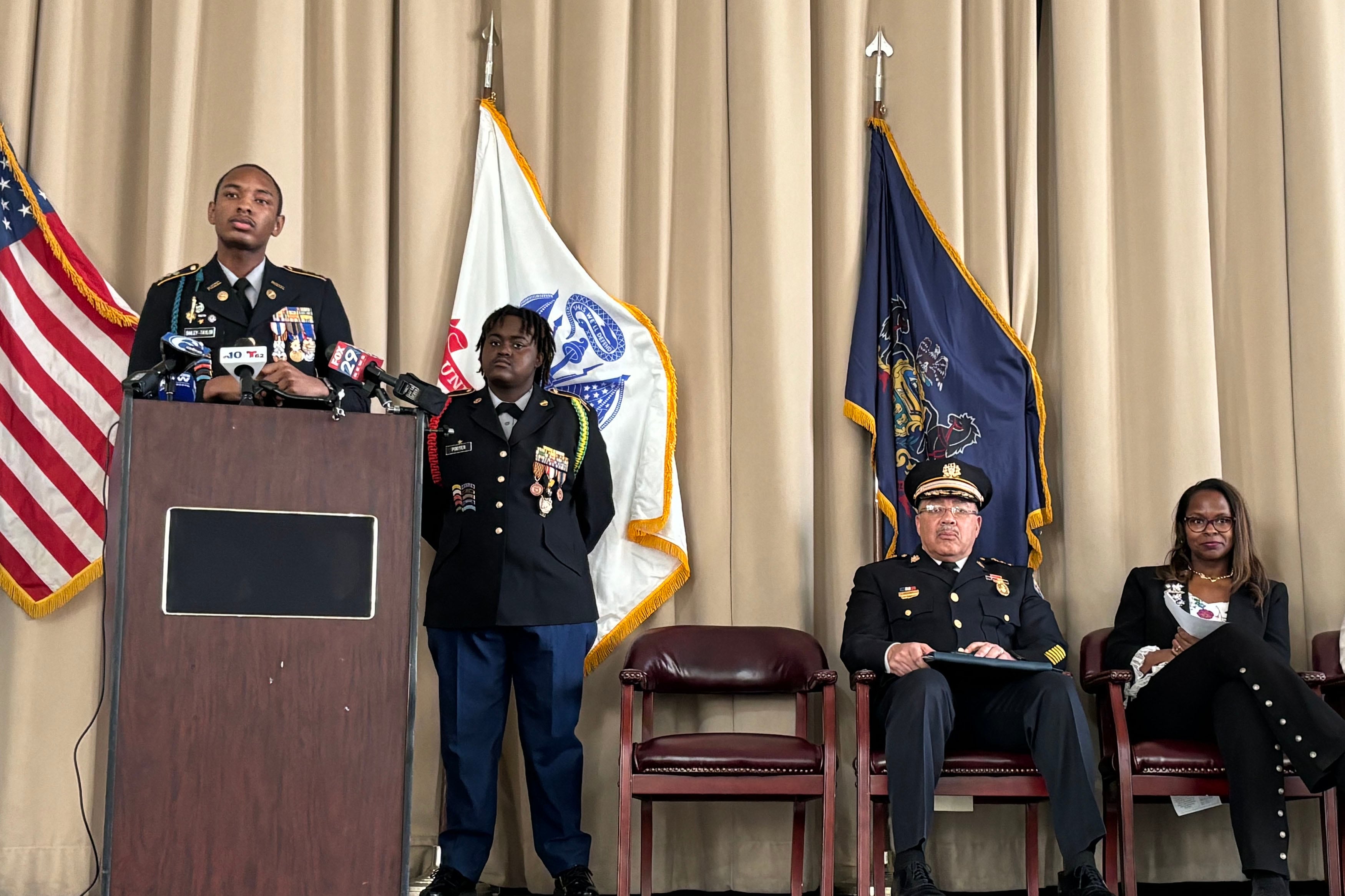 Two students wearing military like uniforms stand on the left of the frame behind a wooden podium while two adults sit in chairs on the right of the frame with three flags in the background.