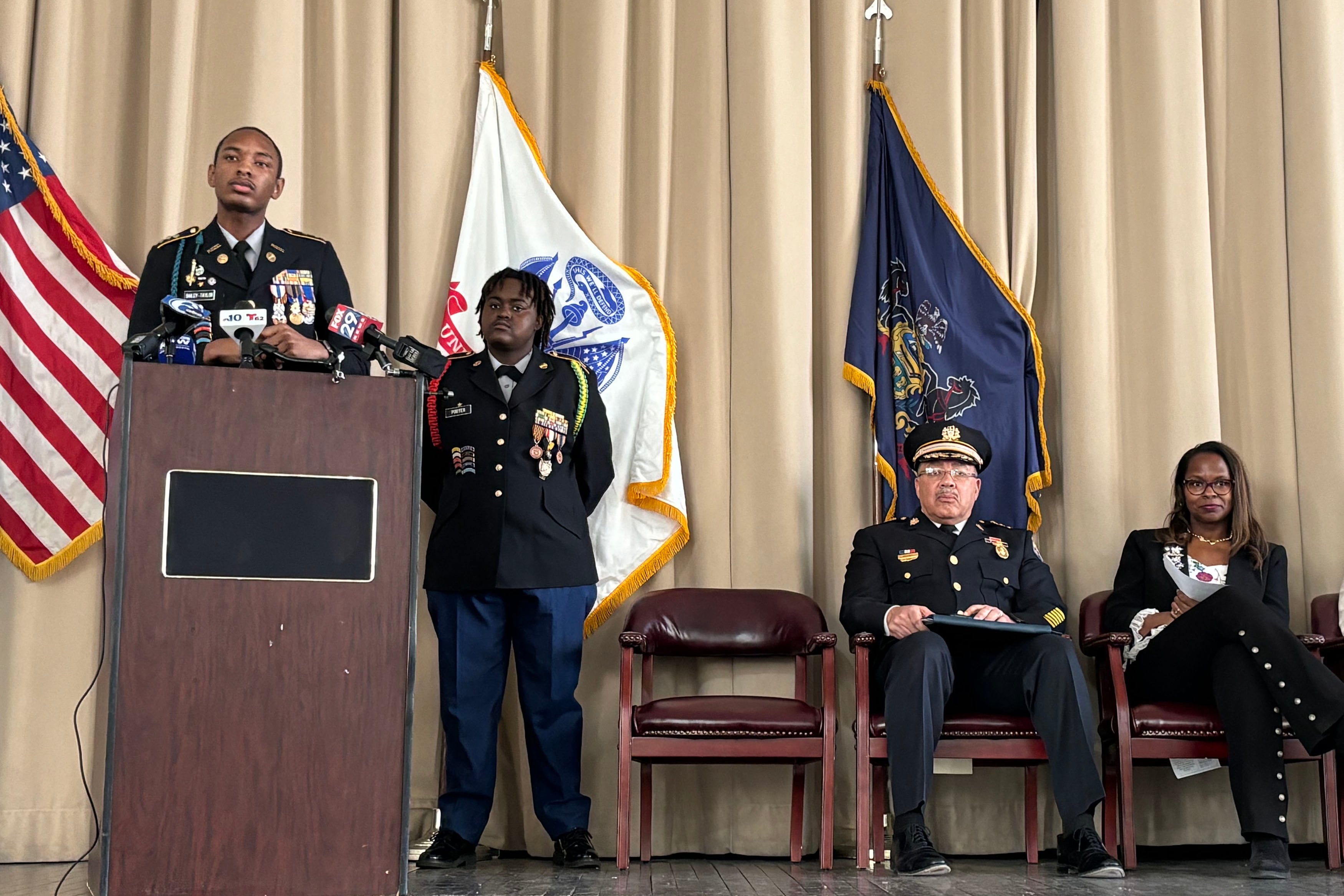 Two students wearing military like uniforms stand on the left of the frame behind a wooden podium while two adults sit in chairs on the right of the frame with three flags in the background.