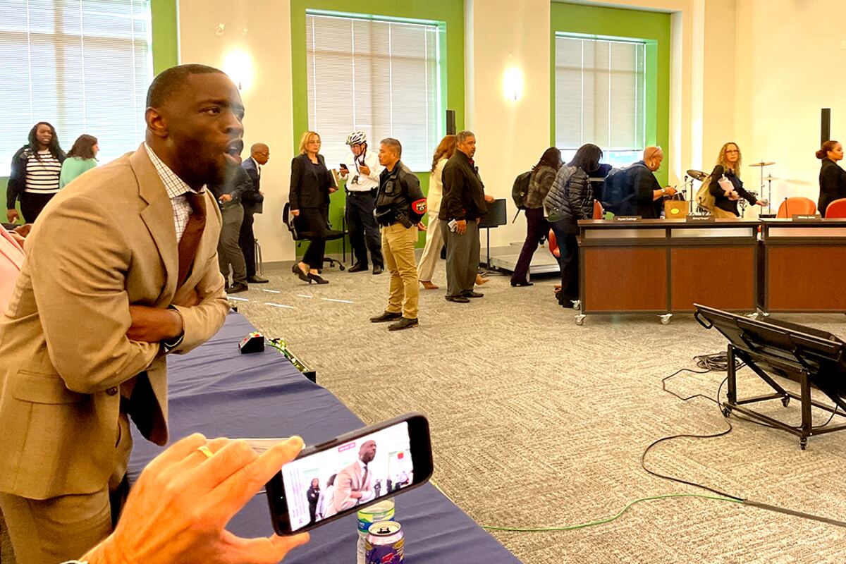 A photograph of a Black man in a light colored suit stands up and speaks passionately during a school board meeting in a room full of other people.