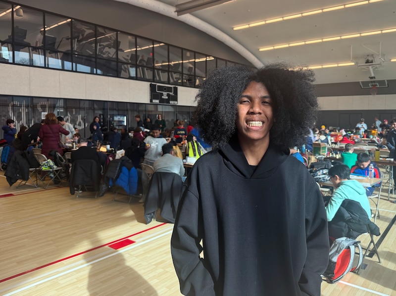 A photograph of a Black high school student posing for a photograph and smiling with a large room in the background with students playing chess.
