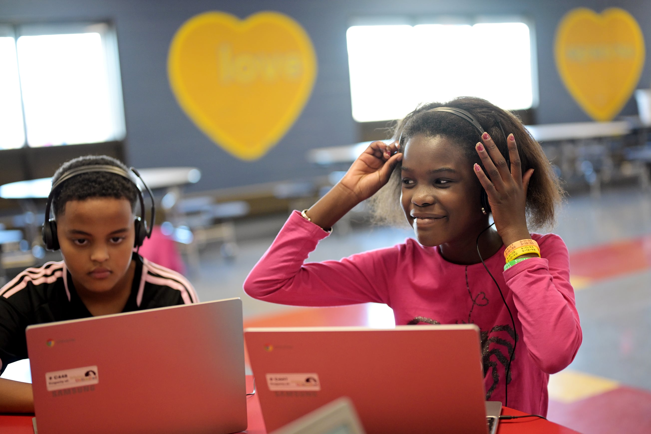 A photograph of two young Black students sitting next to each other at a desk in a colorful classroom each wearing headphones and have a laptop in front of them. One child is looking up and smiling, while the child on the left is reading from the screen.