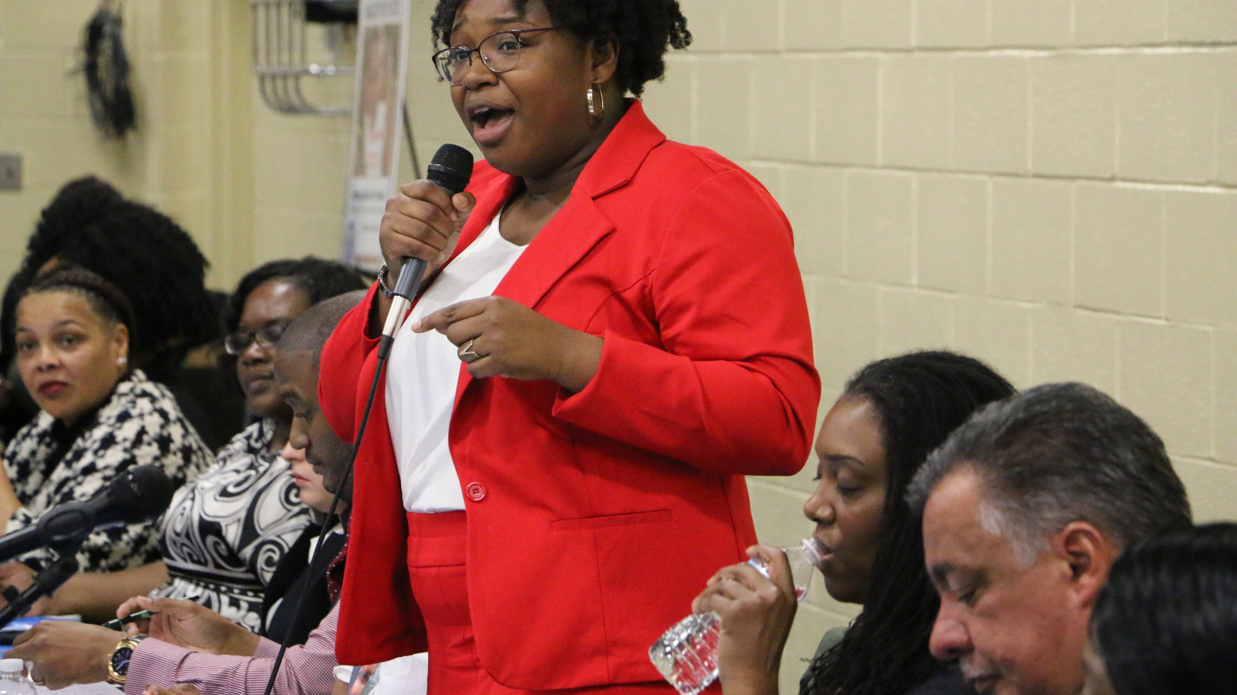 Newark Board of Education member A’Dorian Murray-Thomas. wearing a red suit, speaks while holding a microphone.
