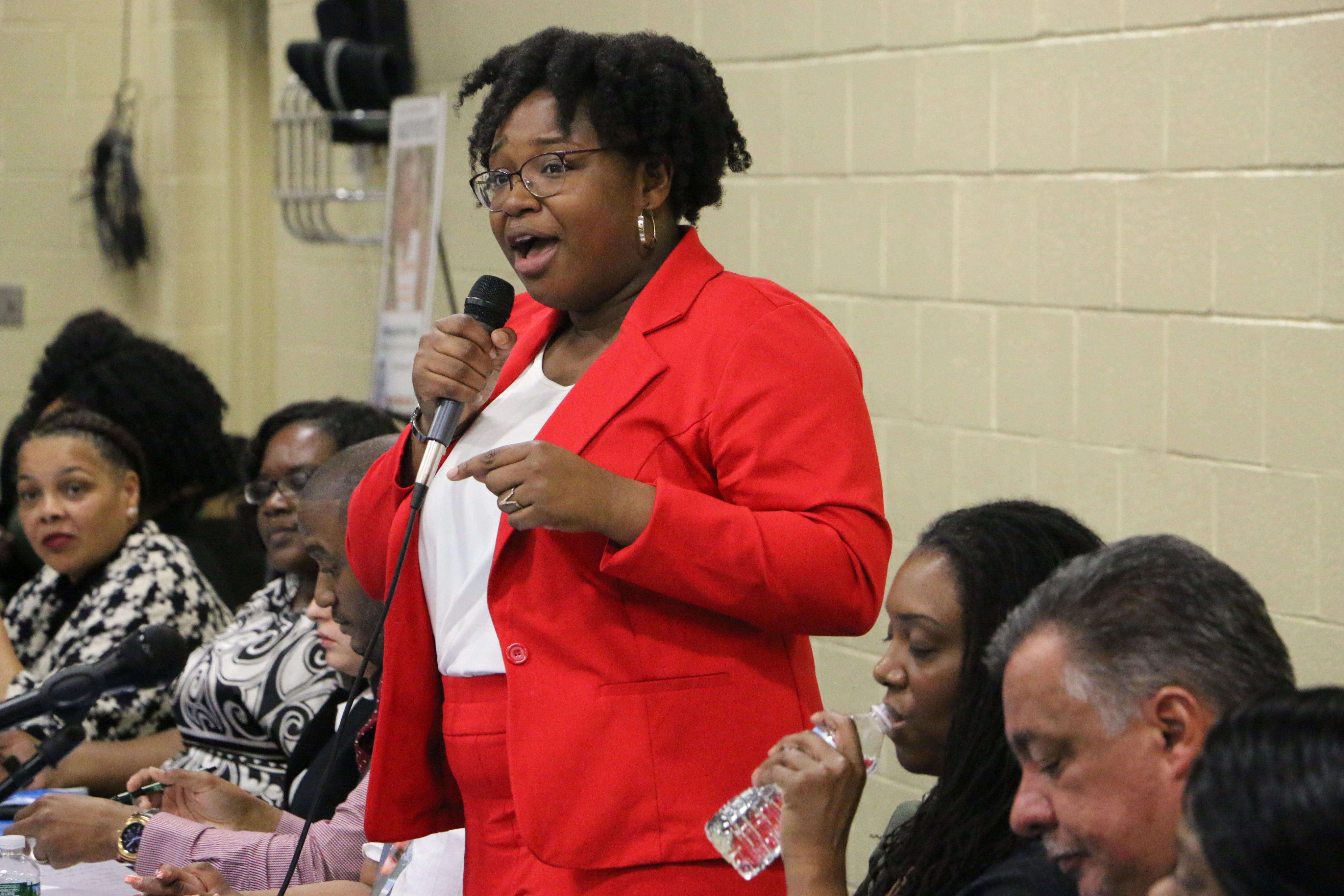 Newark Board of Education member A’Dorian Murray-Thomas. wearing a red suit, speaks while holding a microphone.