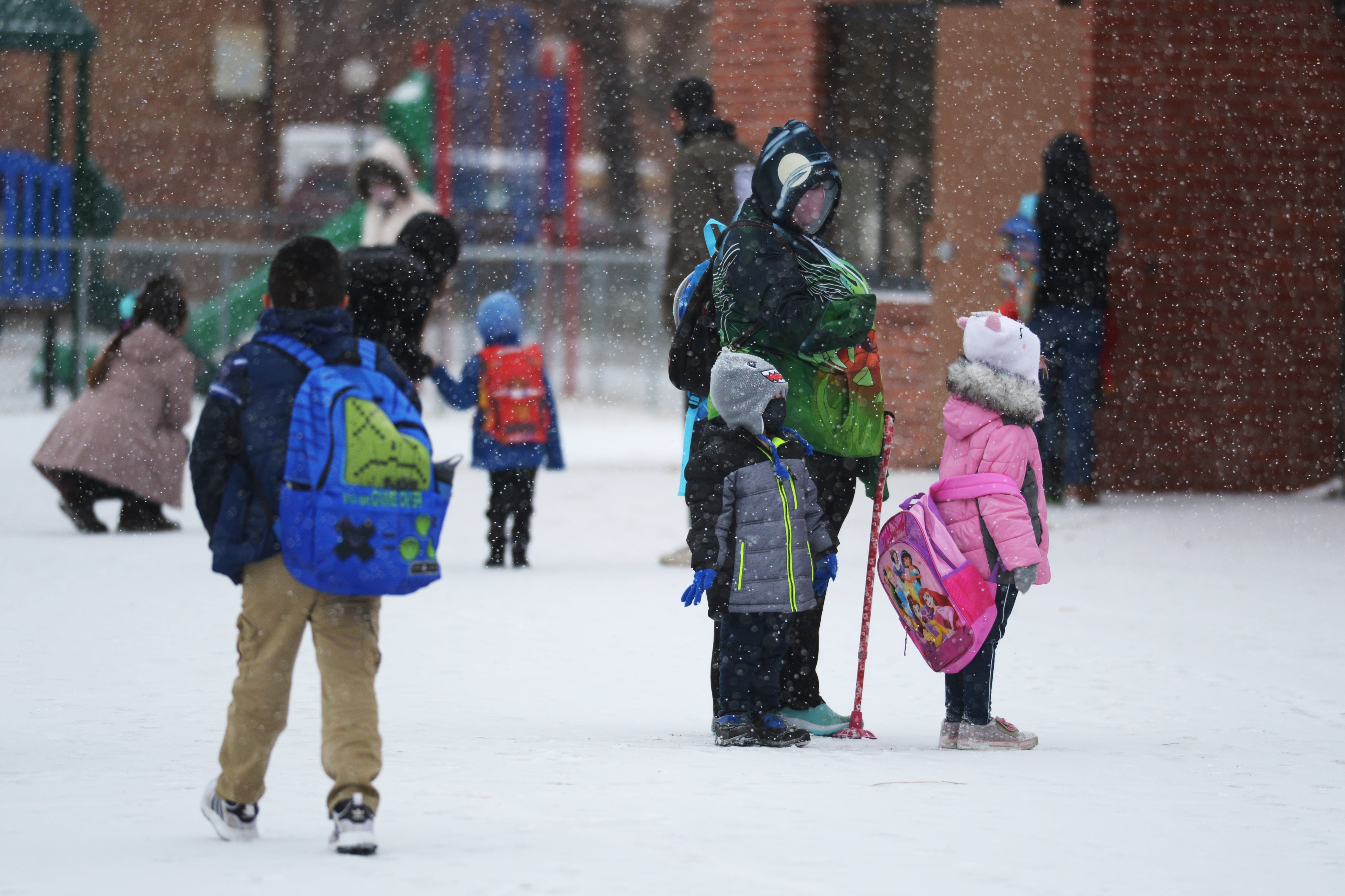 Students and parents wearing masks and winter coats wait outside a school building on a snowy day.