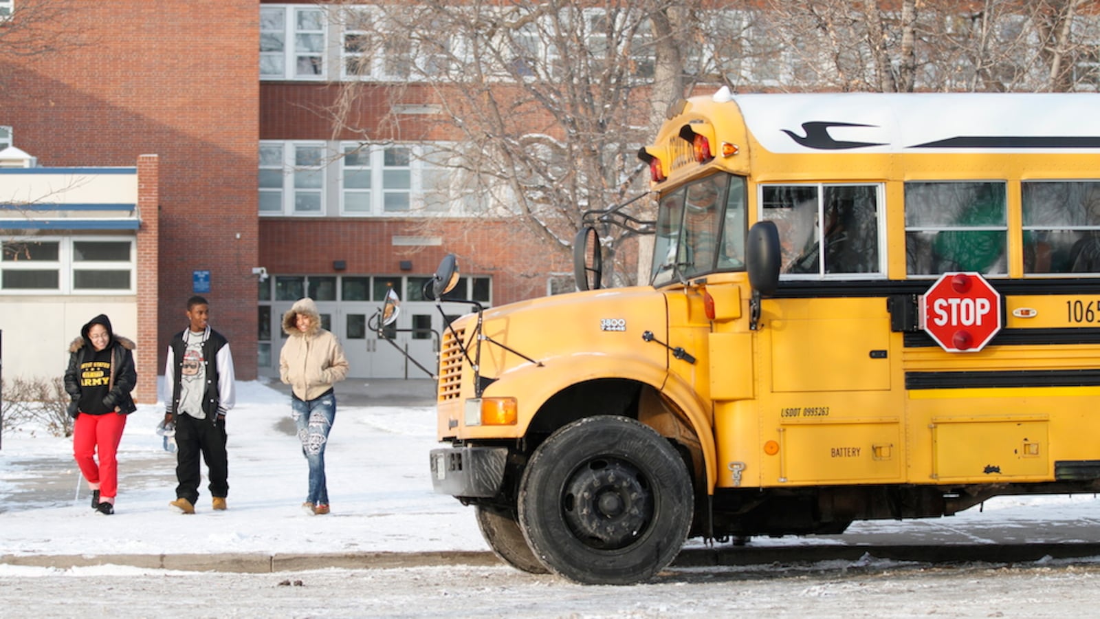 A school bus sits outside a high school in winter. Three students walk by the bus.