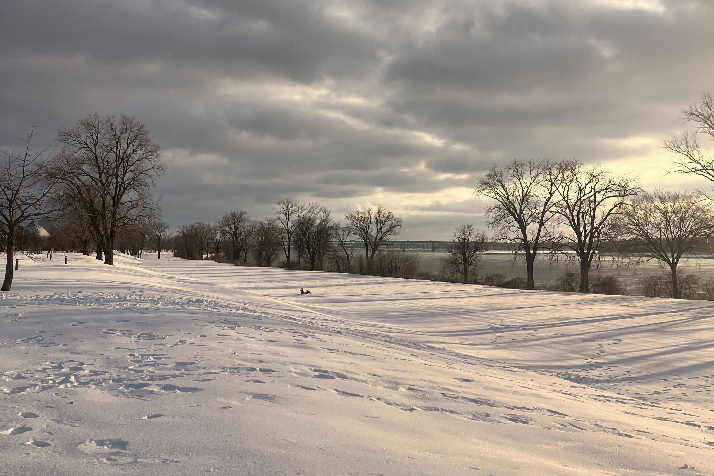 Dark clouds hang over a snow-covered hill will various footprints. Trees line a river in the distance. A single sledder can be spotted on the hill.