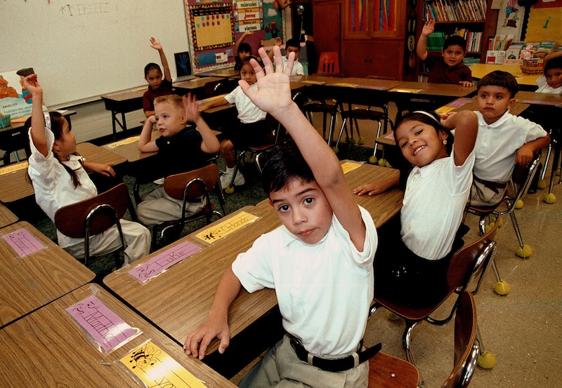 A group of young students all wearing a white shirt and uniform, sit at wooden desks in a classroom. Some of them have their hands up and most are looking directly at the camera.