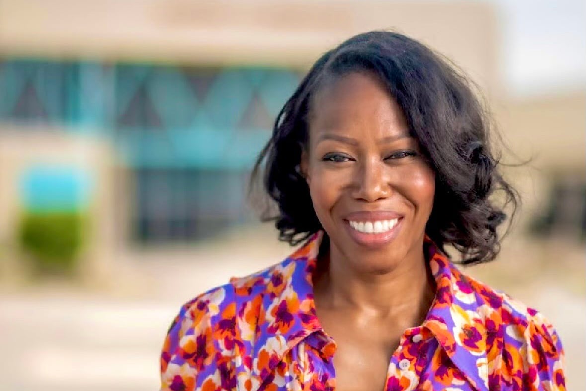 A photograph of a Black woman with short dark hair and wearing a bright blouse smiles while posing for a portrait in front of a school building.