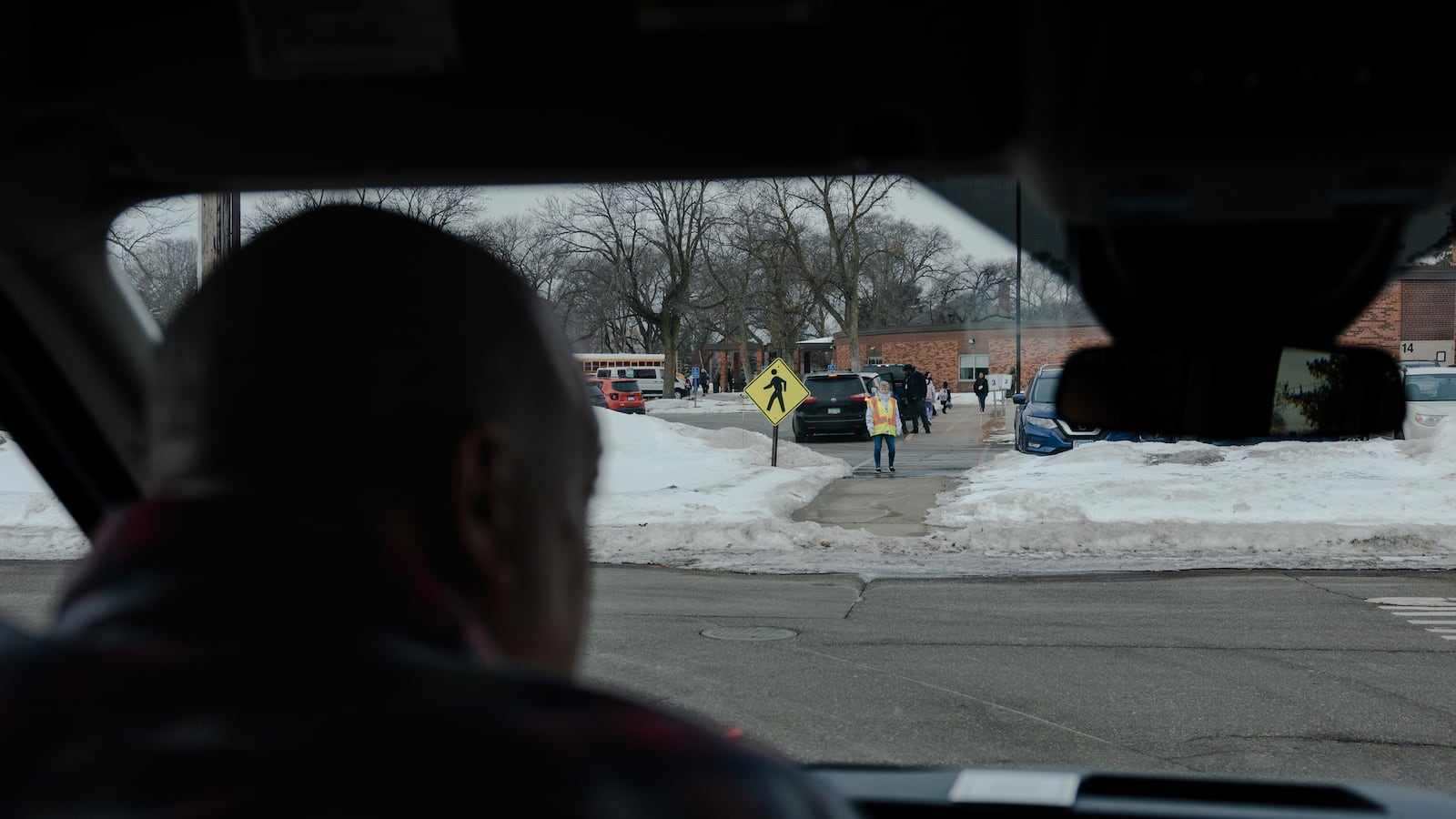 A photograph of a man driving a car while looking out at a crossing guard at a school.