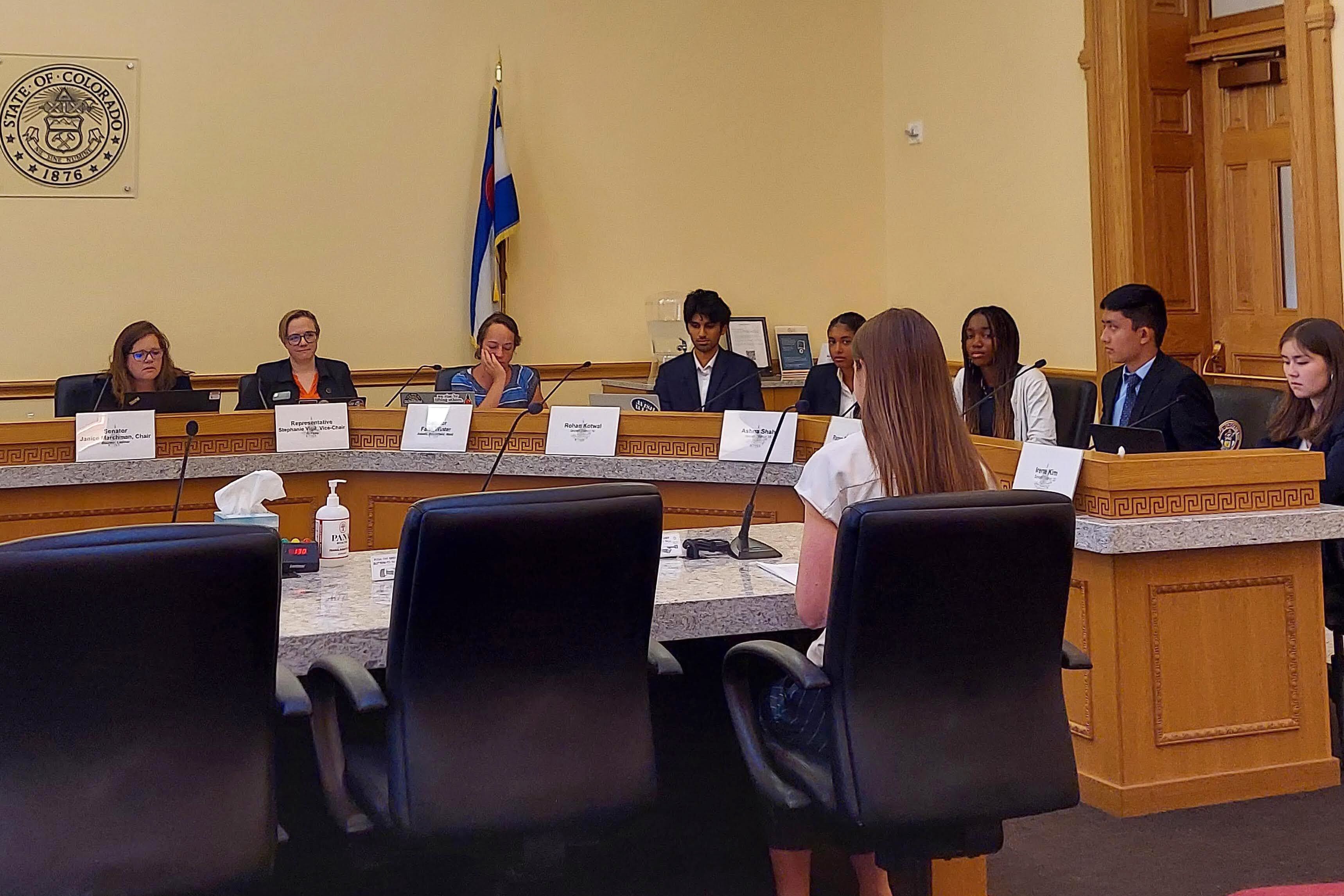 Students and adults sit at chairs around a wooden table in a meeting room.