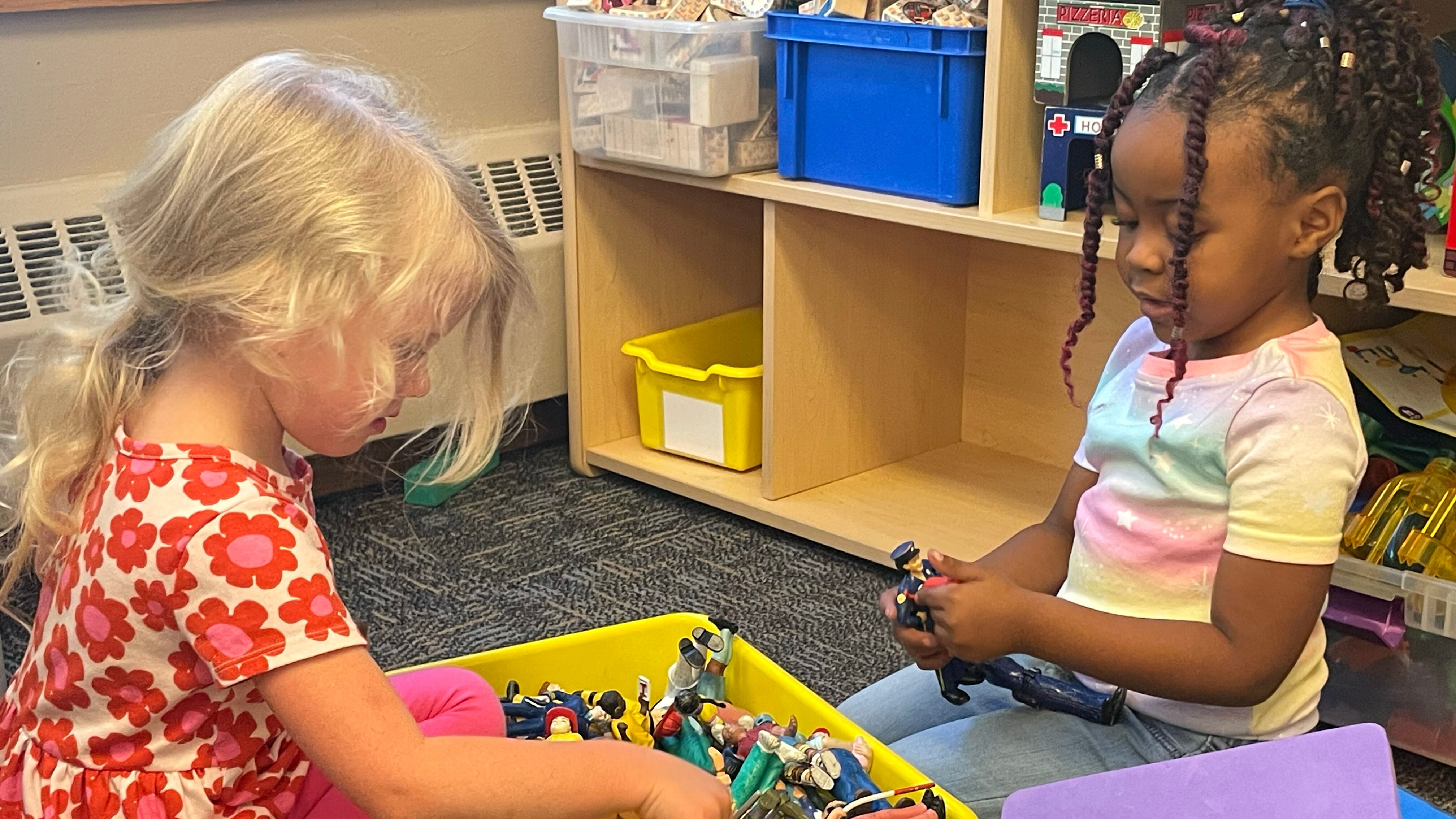 Two preschool girls play with toys from a yellow bin sitting between them.