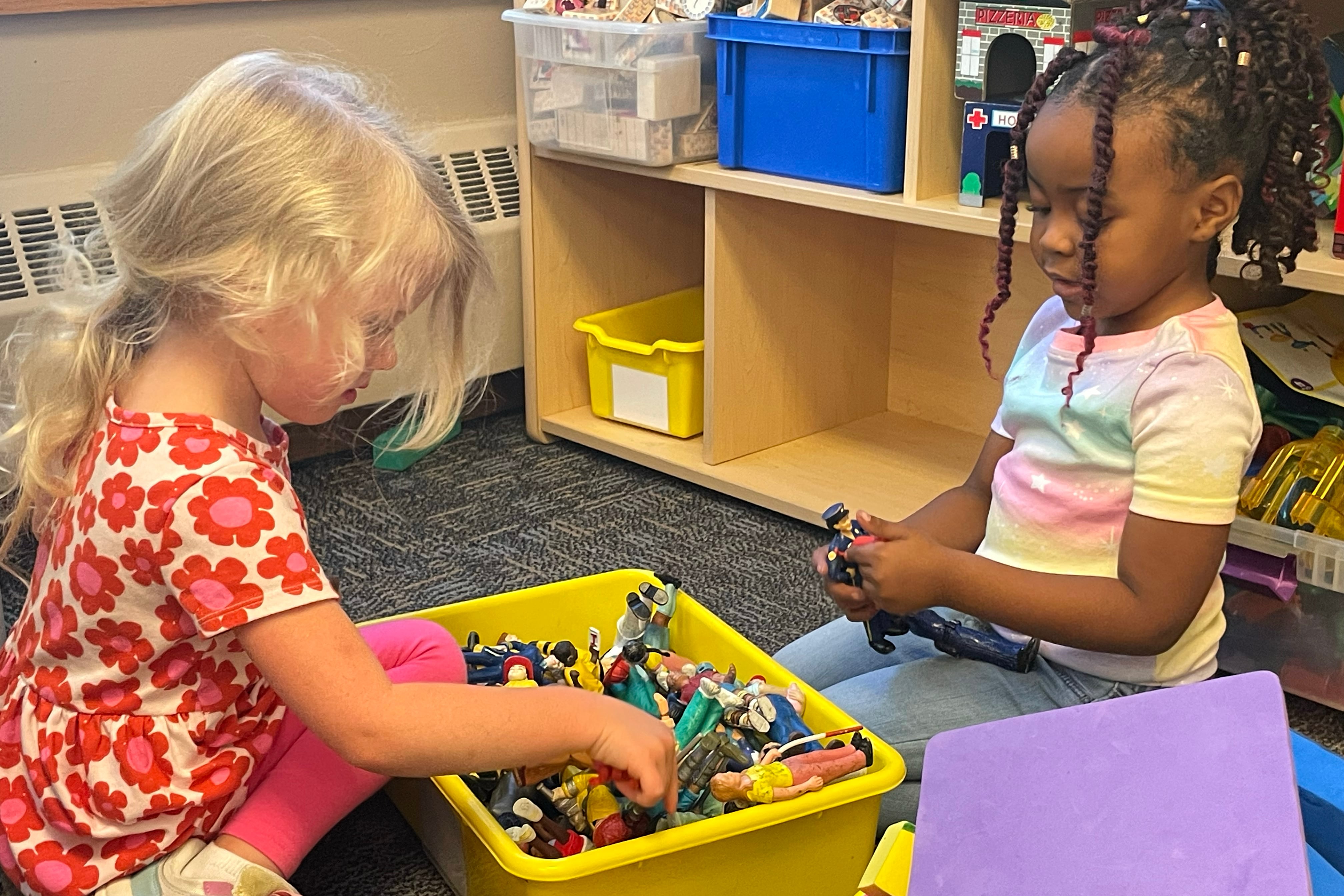 Two preschool girls play with toys from a yellow bin sitting between them.