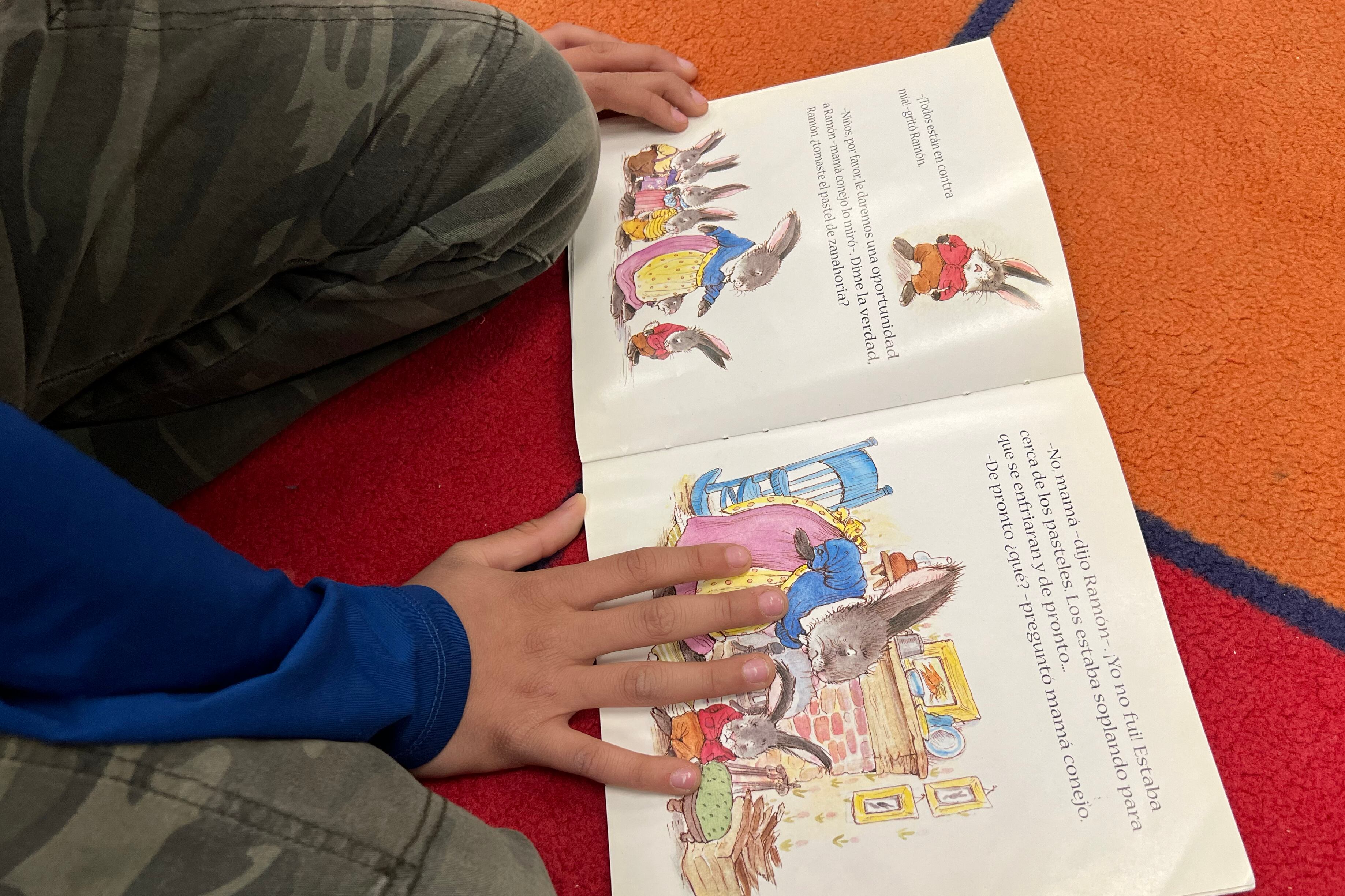Close up of a young student's hands holding down the pages of a book on a colorful rug.