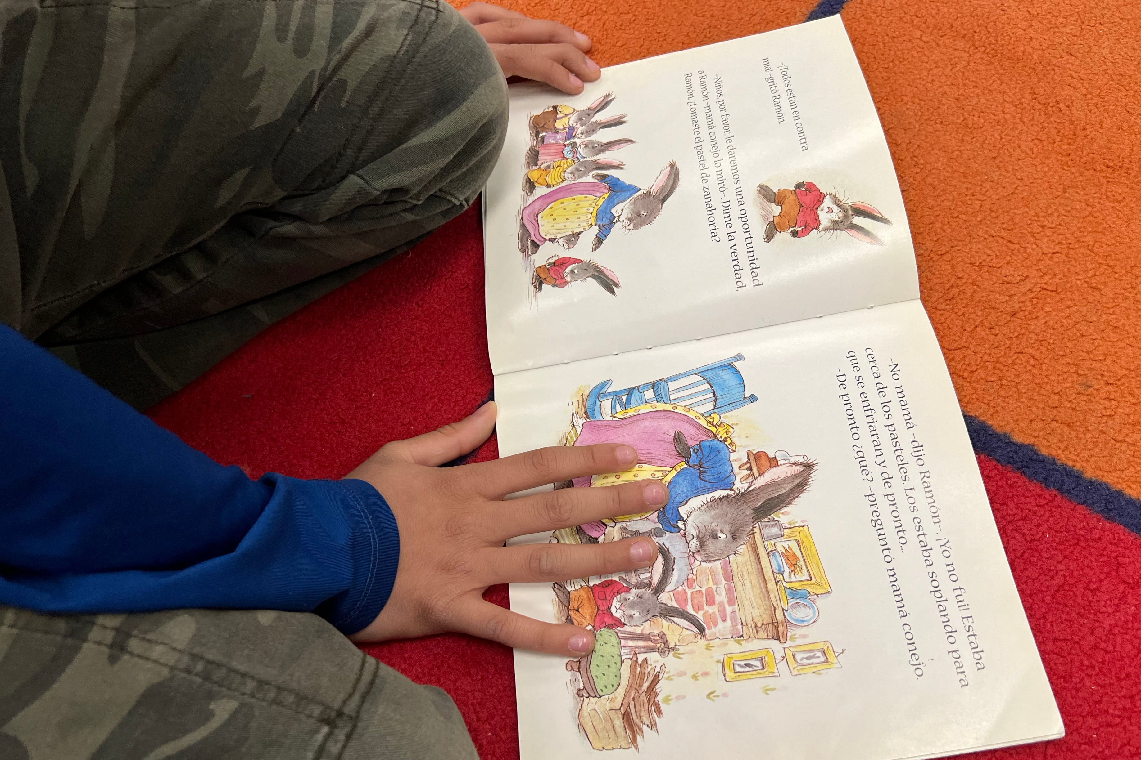 Close up of a young student's hands holding down the pages of a book on a colorful rug.