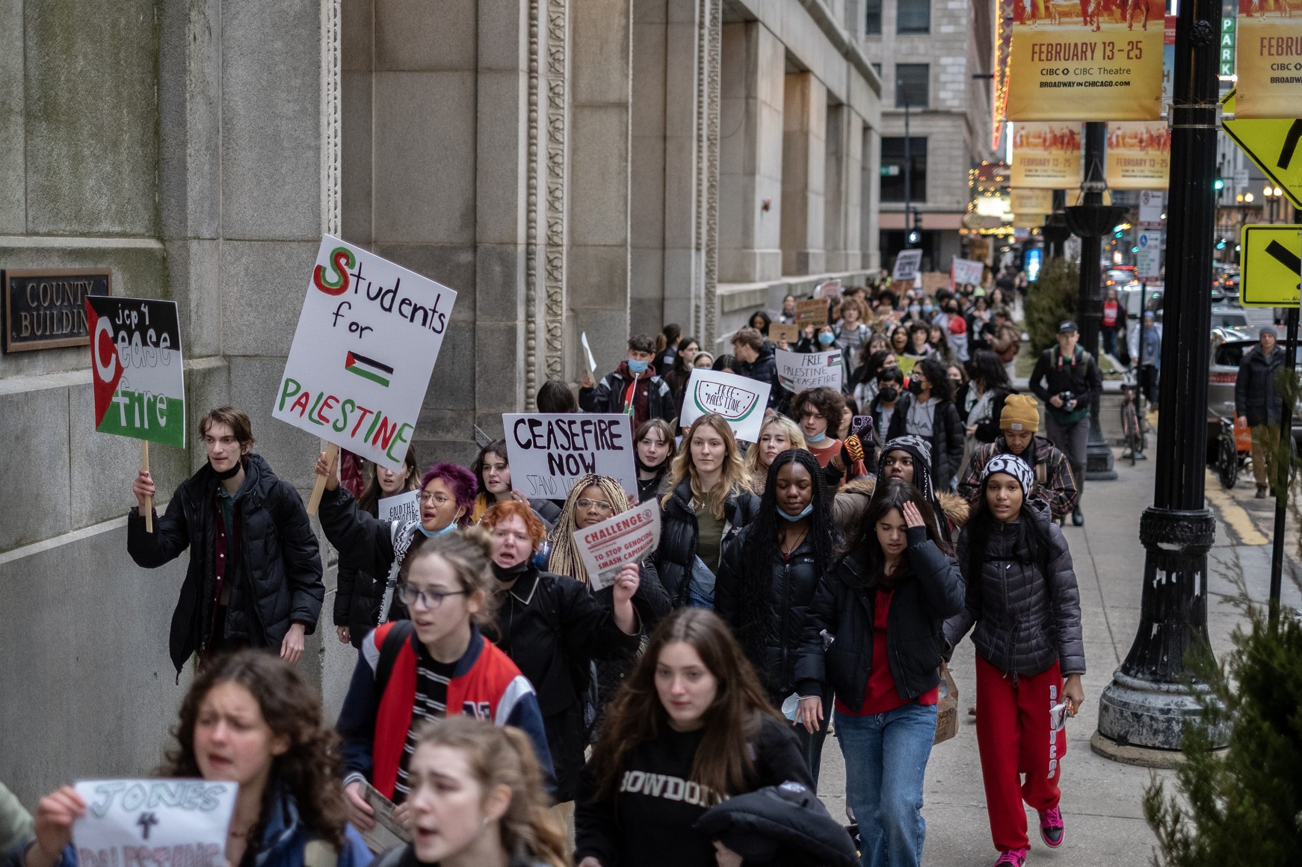A mass of high school students walkout during a protest and march along a sidewalk. Some are holding signs.