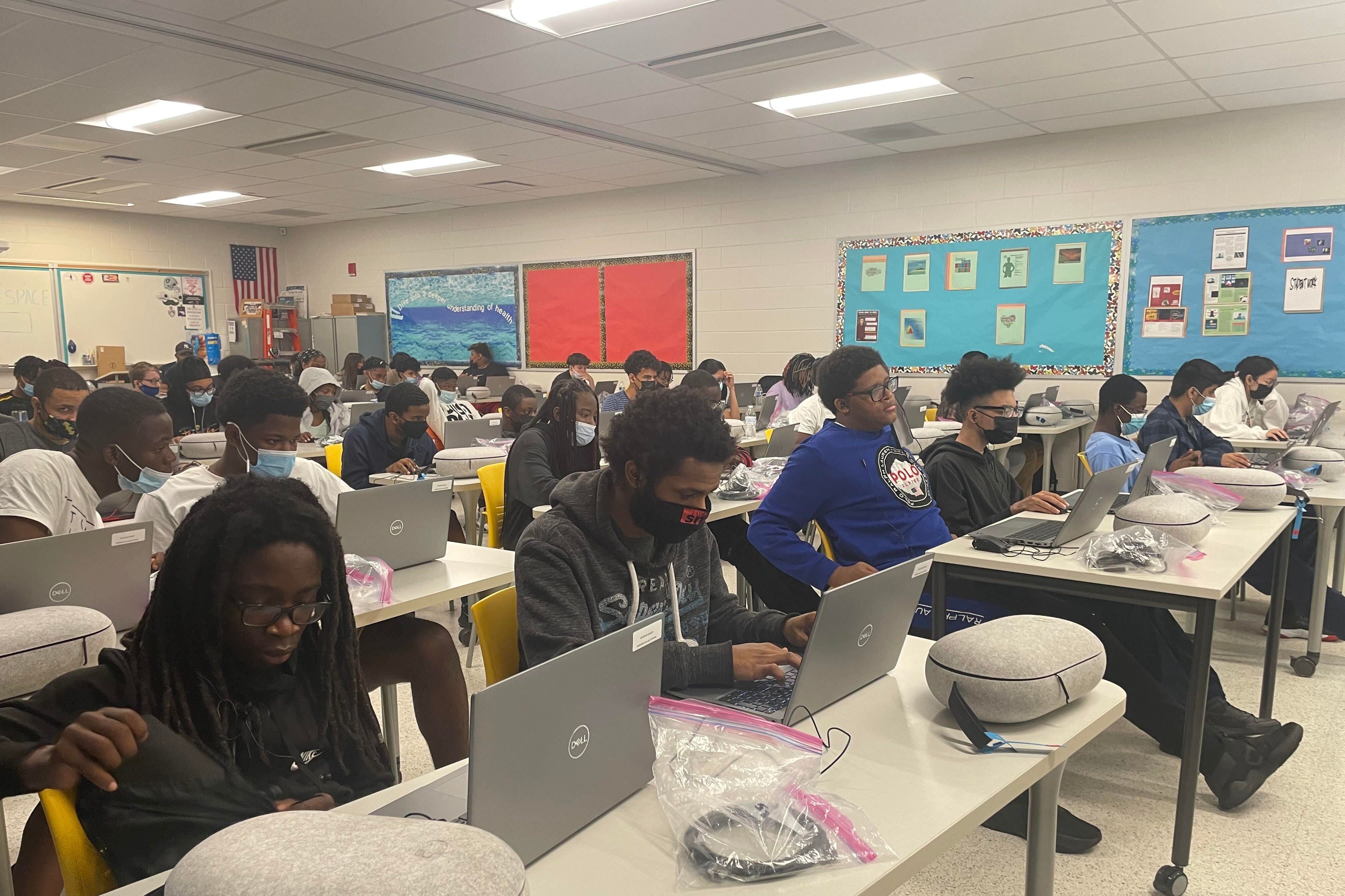 Students sit behind desks in a classroom as they type on their laptops.