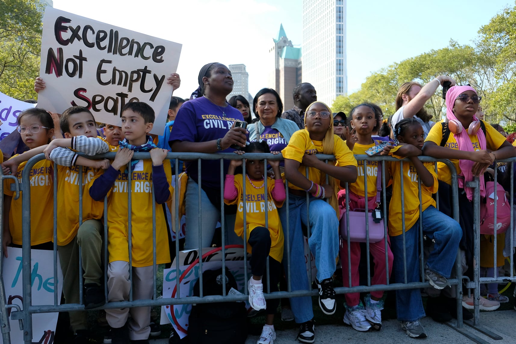 A large group of students and youth, some wearing yellow shirts and some wearing purple, hold signs and wait behind a fence.
