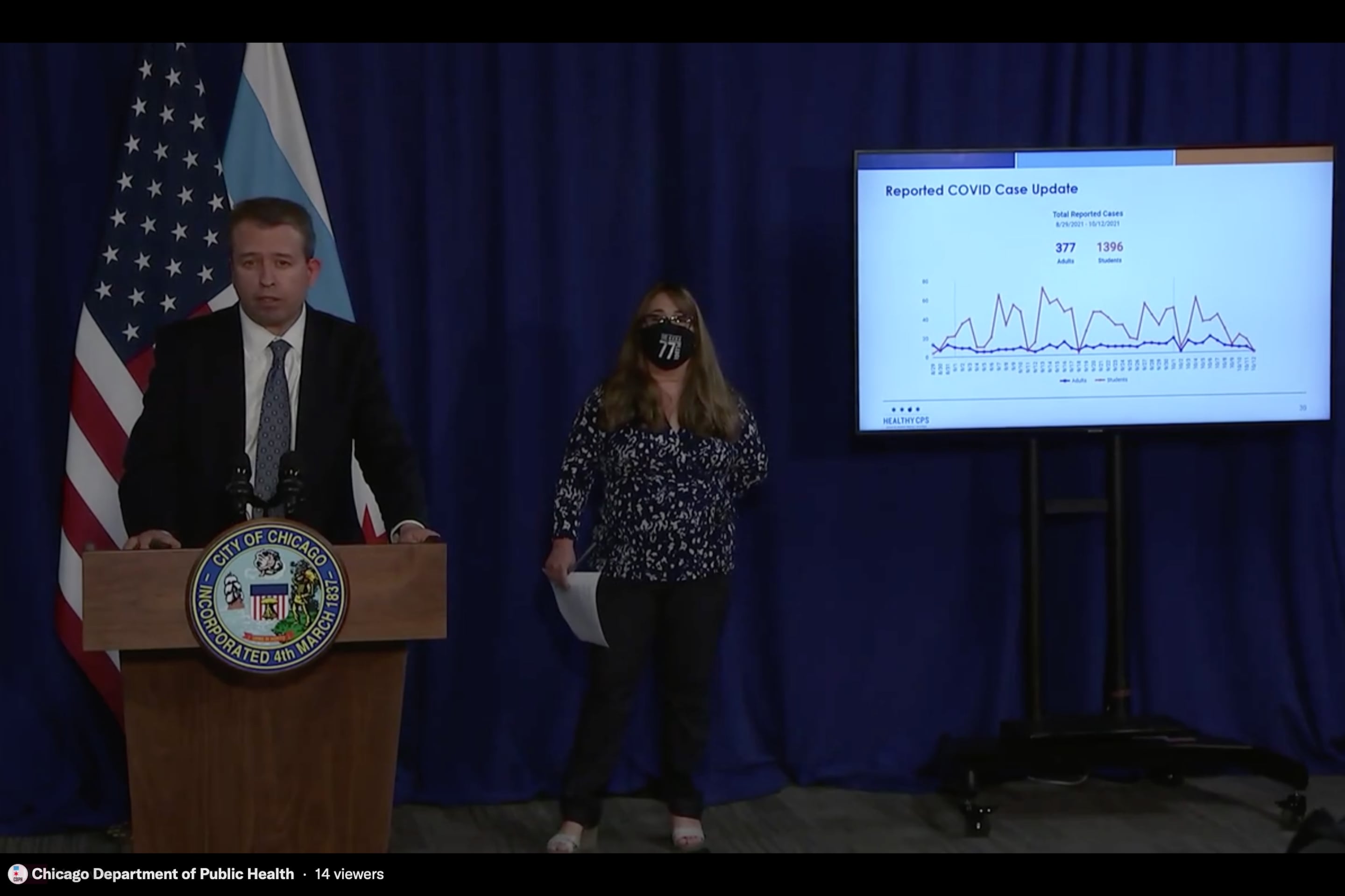 A man in a suit stands beside a sign language interpreter and a screen showing a PowerPoint presentation.