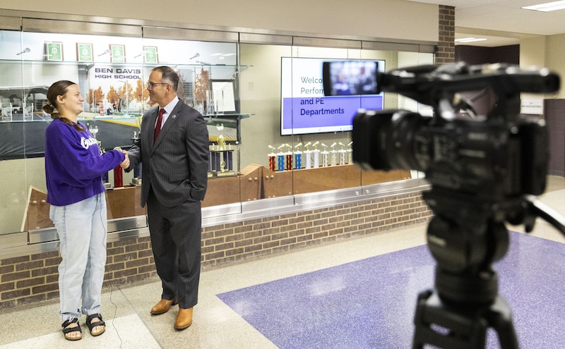 A photograph of a white female student standing next to a white male adult in front of a video camera.