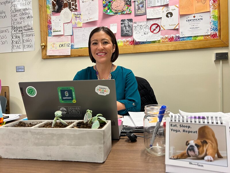 A woman with short dark hair sits behind a laptop and in front of a wall with a cork board on the wall.