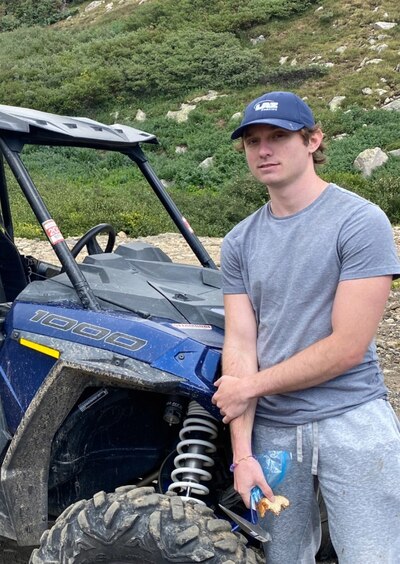 A person wearing a blue hat and a grey shirt holds a half-eaten sandwich and standing next to an off-road vehicle.