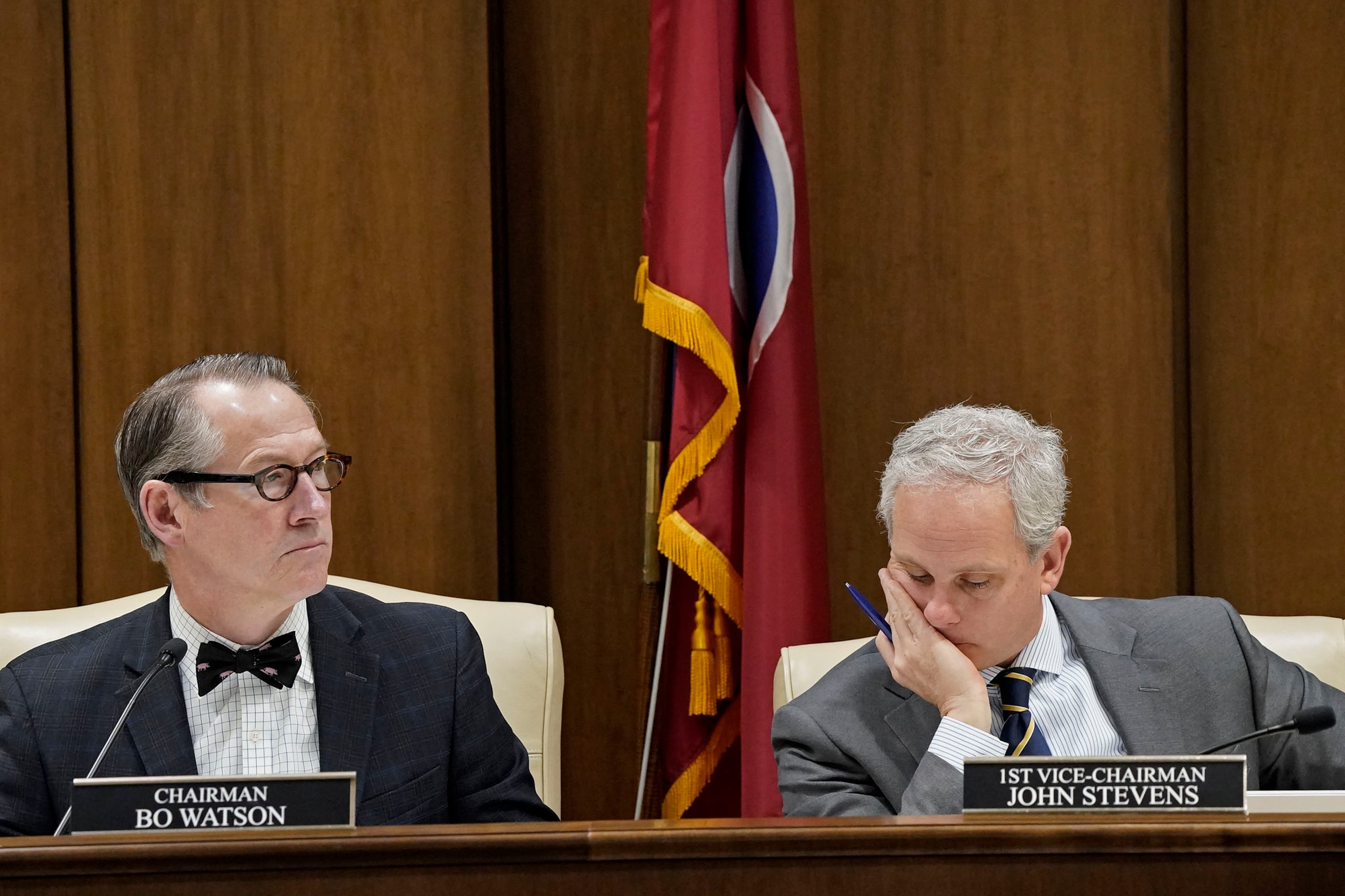 Two men wearing suits sit at a wooden table with a wooden wall and a flag in the background.