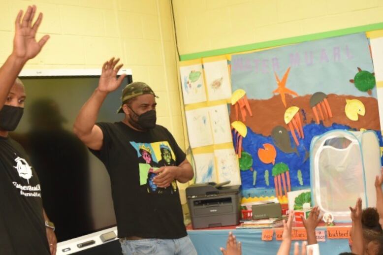 Two men stand in front of a screen with their right hands raised in the air while wearing black T-shirts and masks.