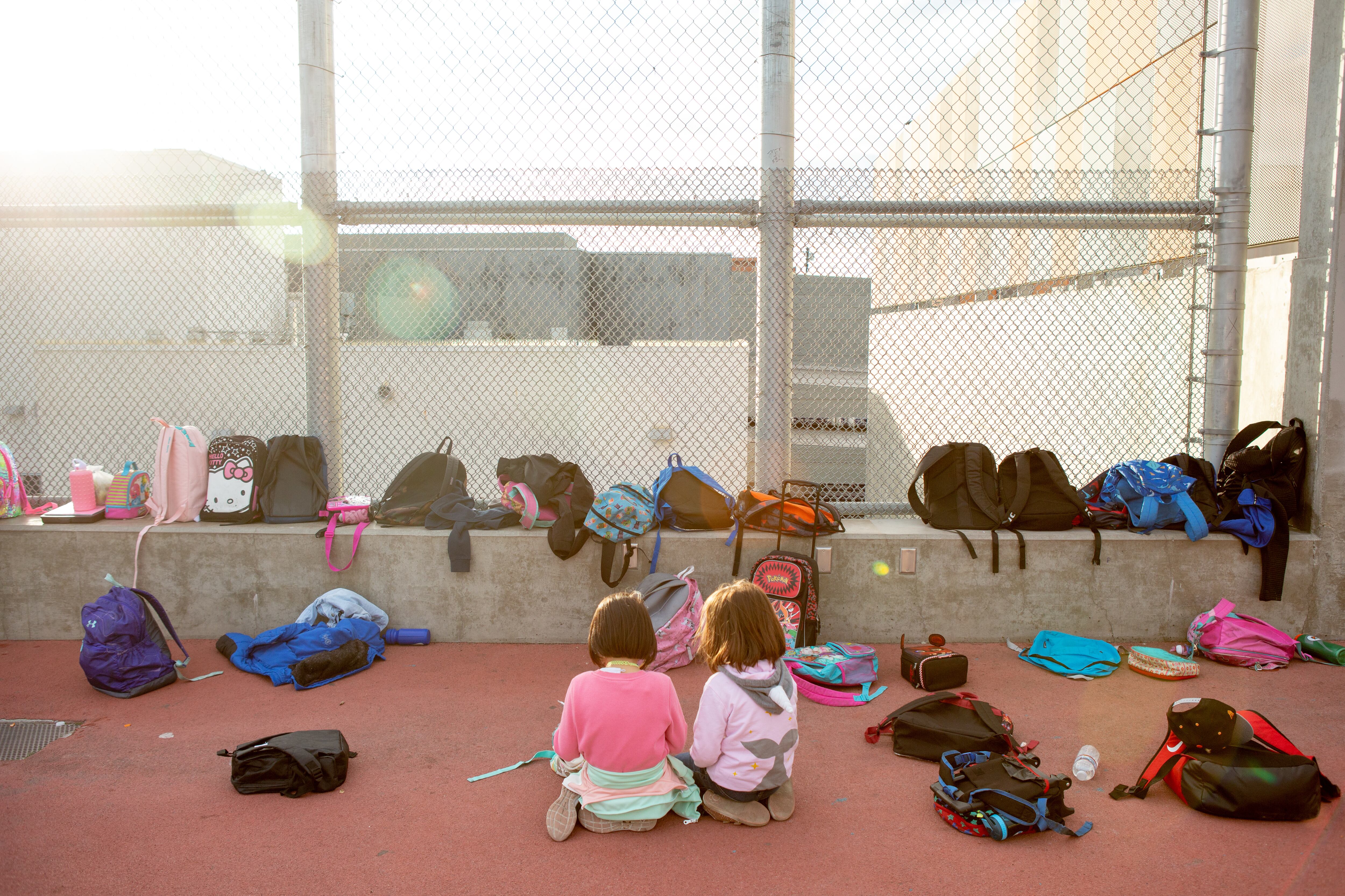 Two young students sit outside next to more than a dozen backpacks.