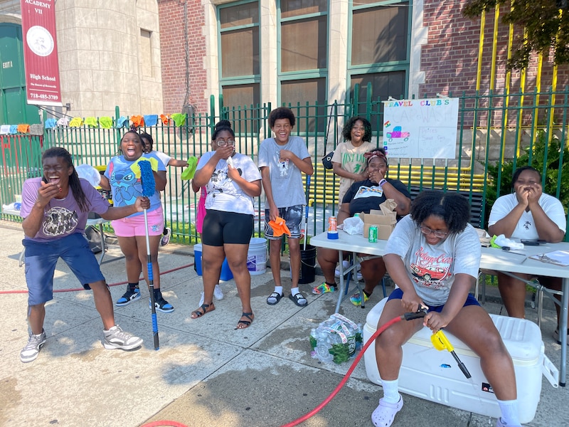 A group of students outside, some are standing and some are sitting when the sun is shining on a large brick school building in the background.
