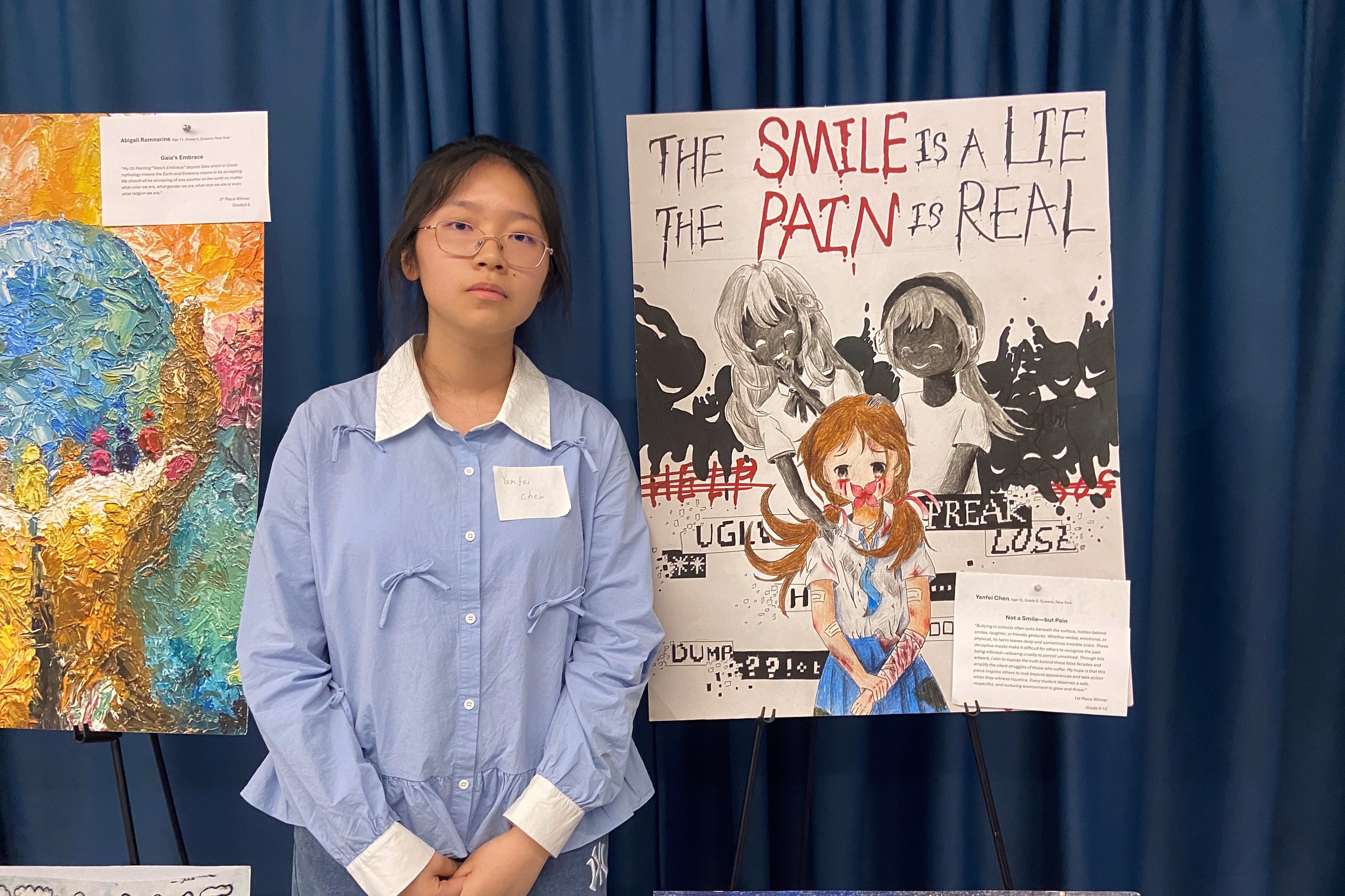 A photograph of a student with dark hair and wearing glasses standing next to a large piece of artwork.