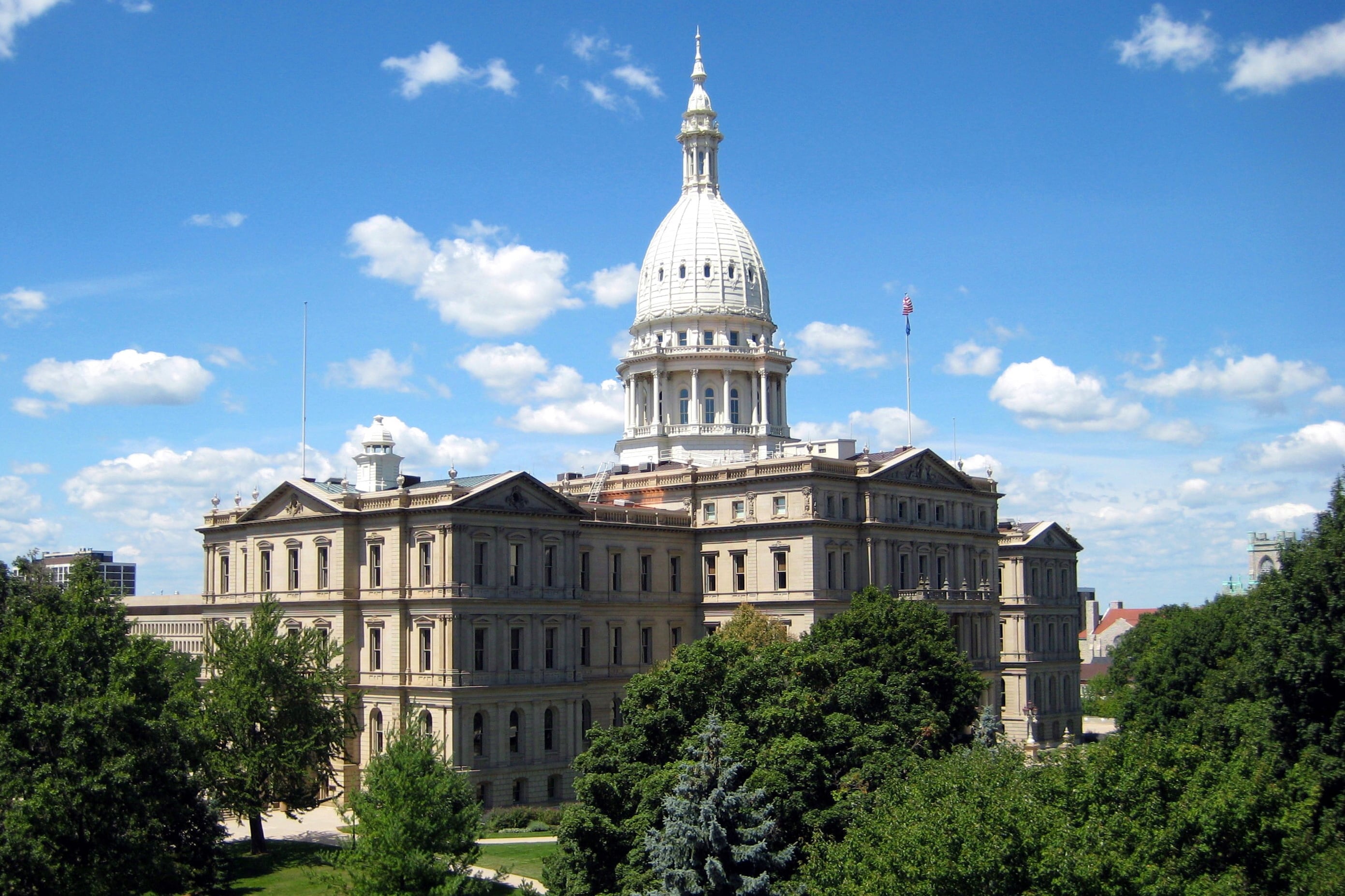 An aerial view of the Michigan State Capitol building in the summer surrounded by blue skies and lush, green trees.