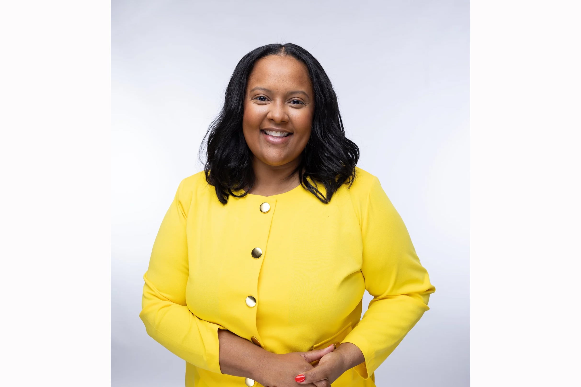 A Black woman in a bright yellow suit and medium length dark hair smiles while posing for a portrait against a grey backdrop.