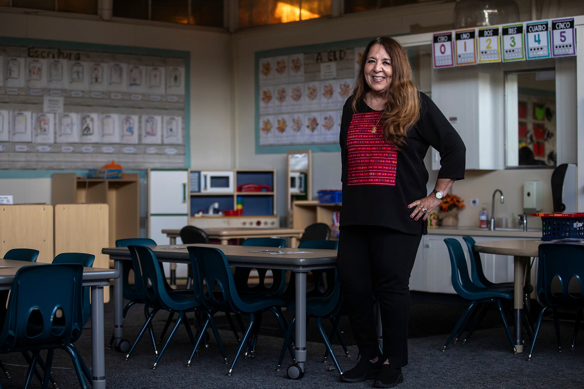 A woman with a red shirt and black jacket and pants stands in an empty classroom.