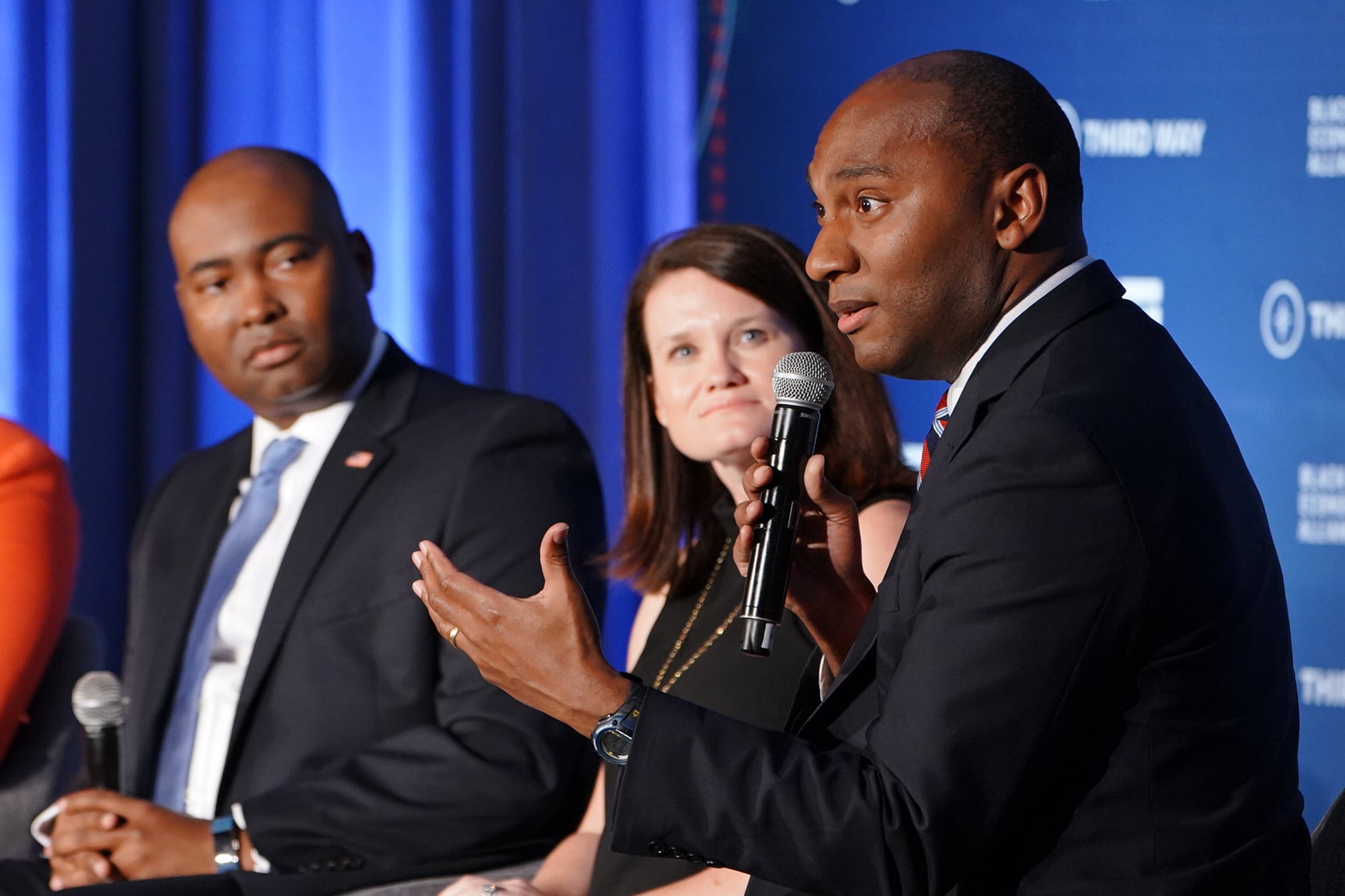 A photograph of a Black man in a suit speaking from a microphone with two others in the background.