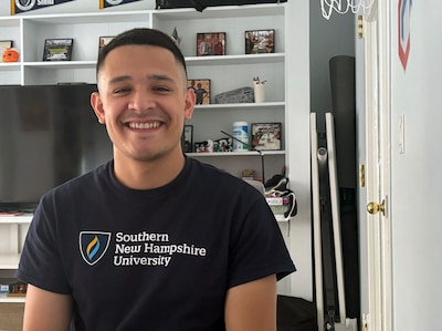 A high school senior with short dark hair and wearing a black t-shirt poses for a portrait in front of a bookshelf.