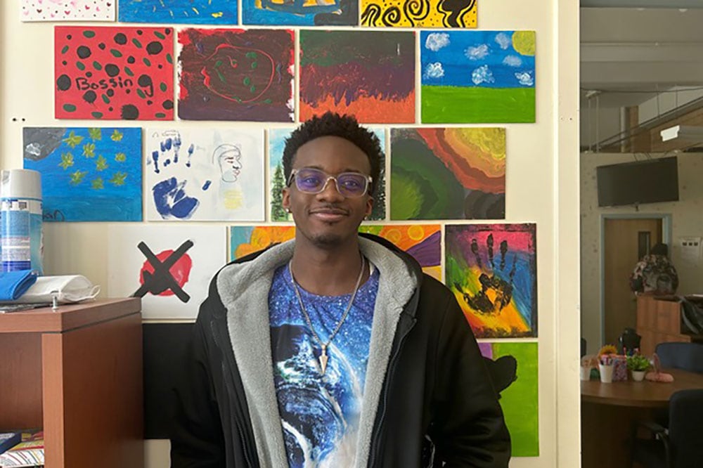 A young Black man with short hair and wearing a black jacket over a blue shirt poses for a portrait while smiling and standing in front of a wall covered in art.