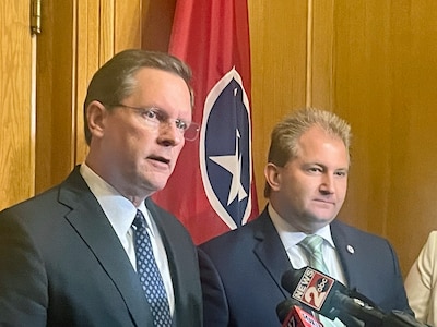Two men wearing suits stand next to each other with a flag in the background.