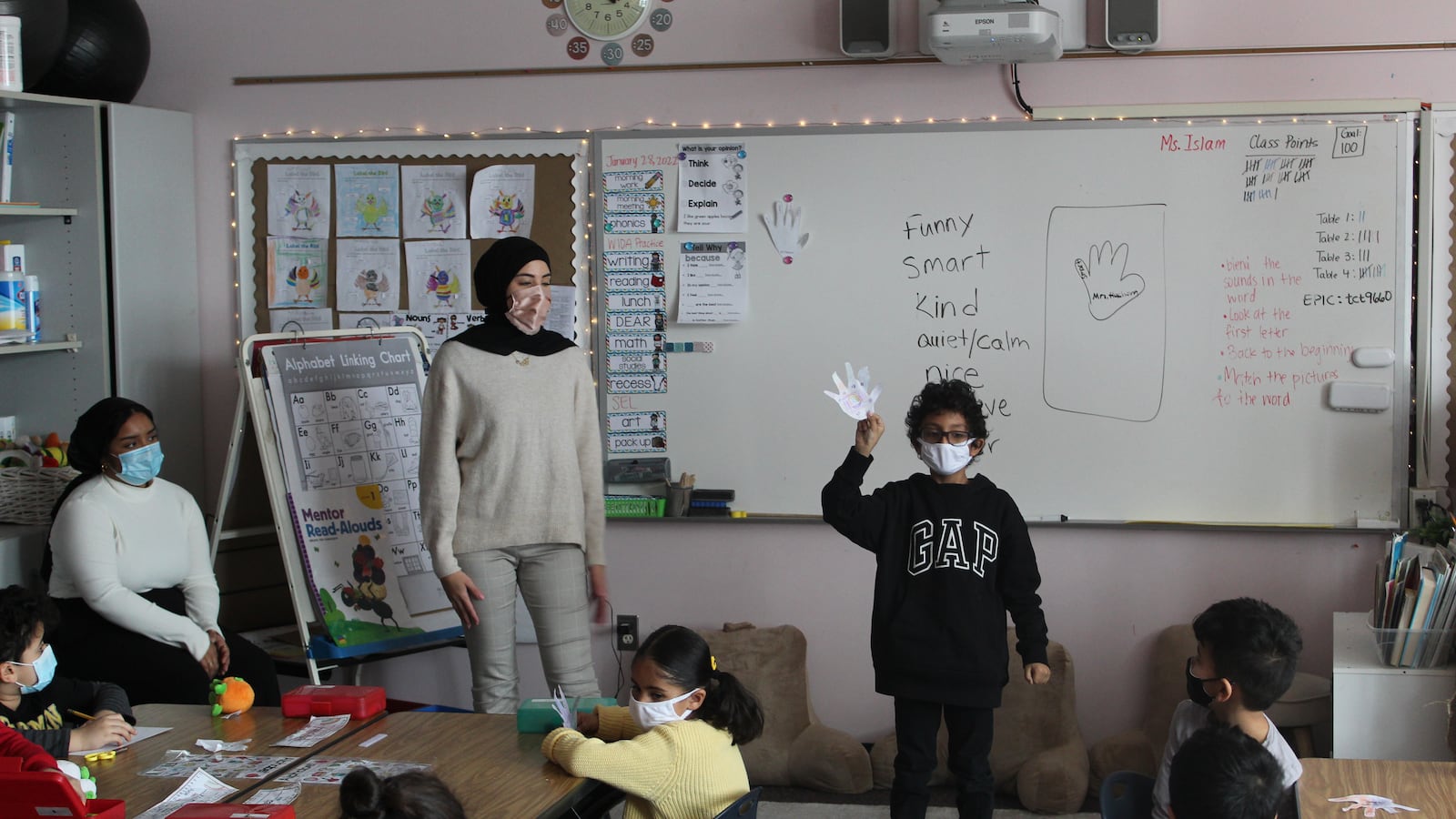 a child in black sweater holds up a paper hand standing next to a teacher at the front of the classroom