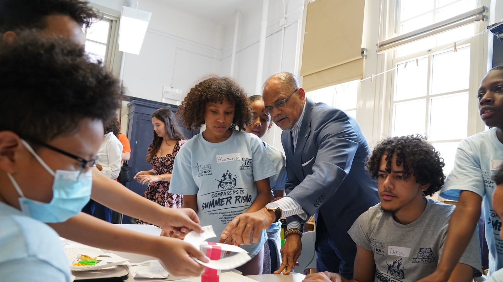 A man in a suit stands with students in T-shirts in a classroom.