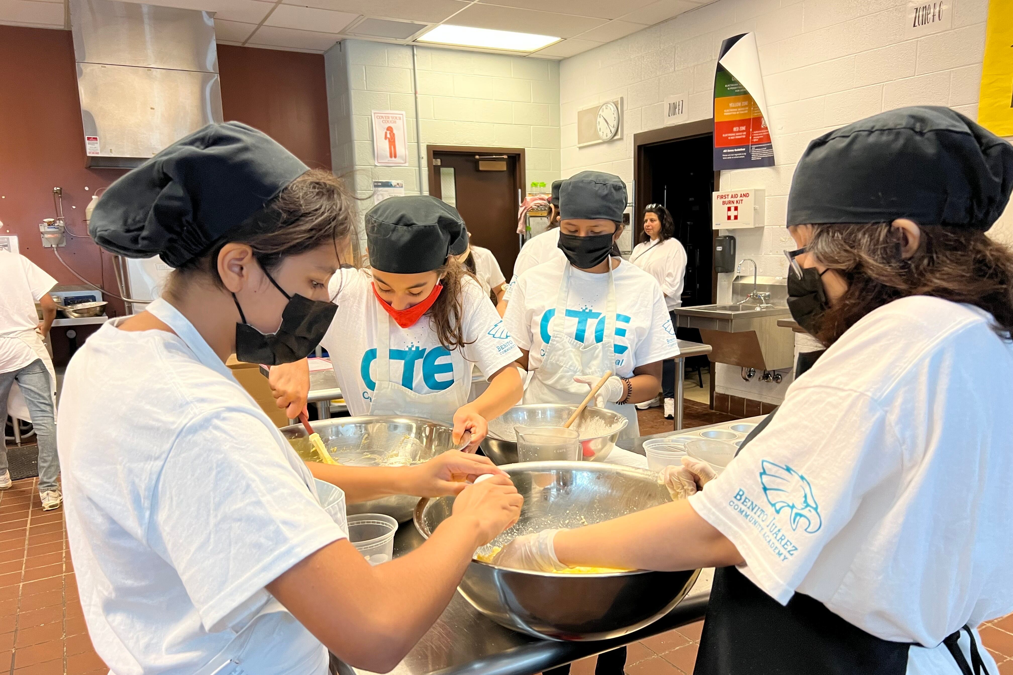 Four students in white camp t-shirts and chef caps gather around a table mixing cupcake batter in a large bowl.