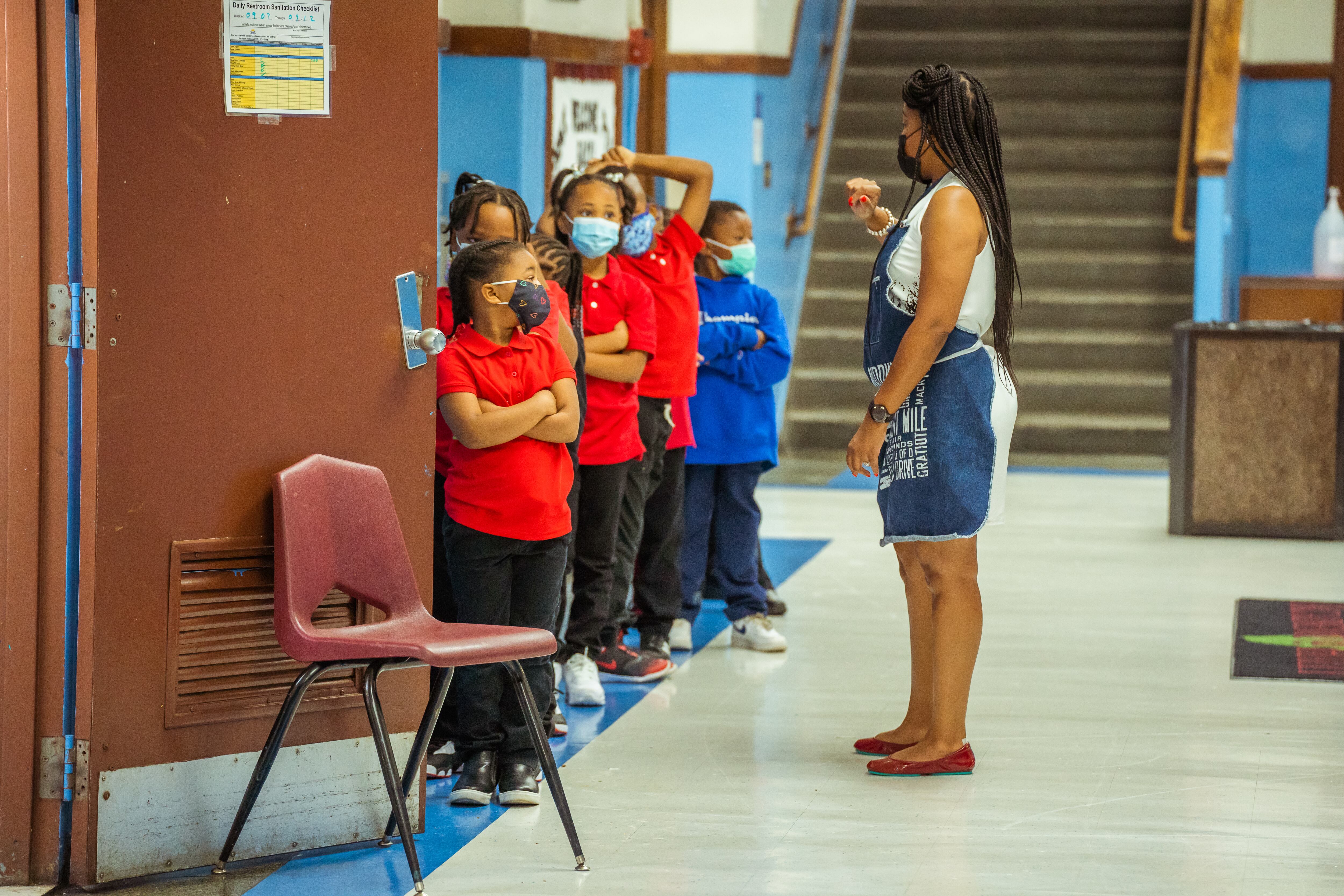 Students form in single-file line as a teacher provides instructions in a school hallway.