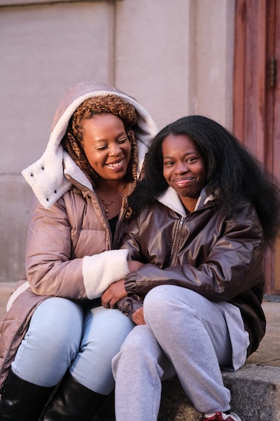 An adult woman and her daughter wear winter jackets and sit next to each other posing for a portrait outside.