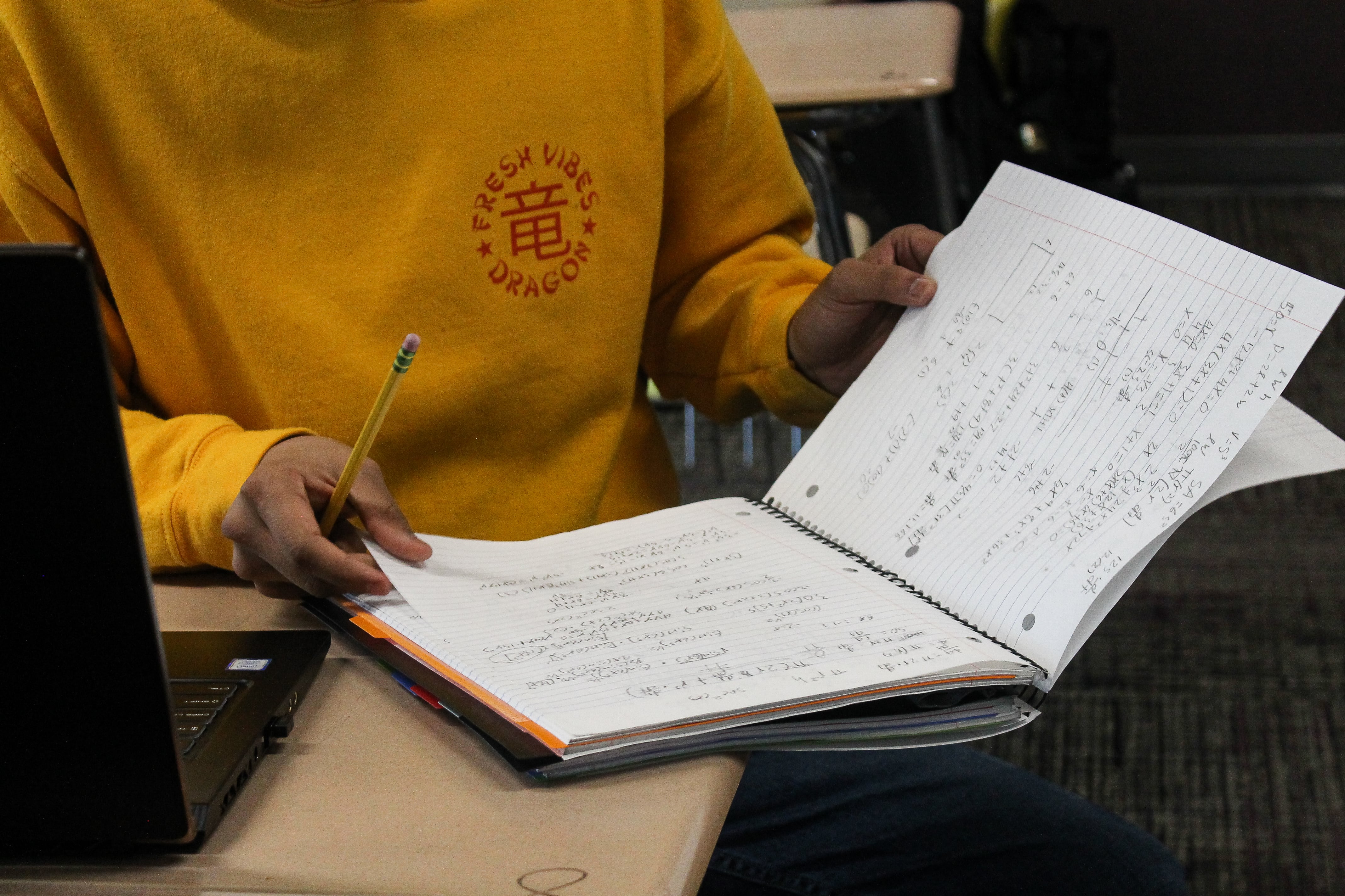 A student in a yellow sweatshirt opens a notebook at their desk.