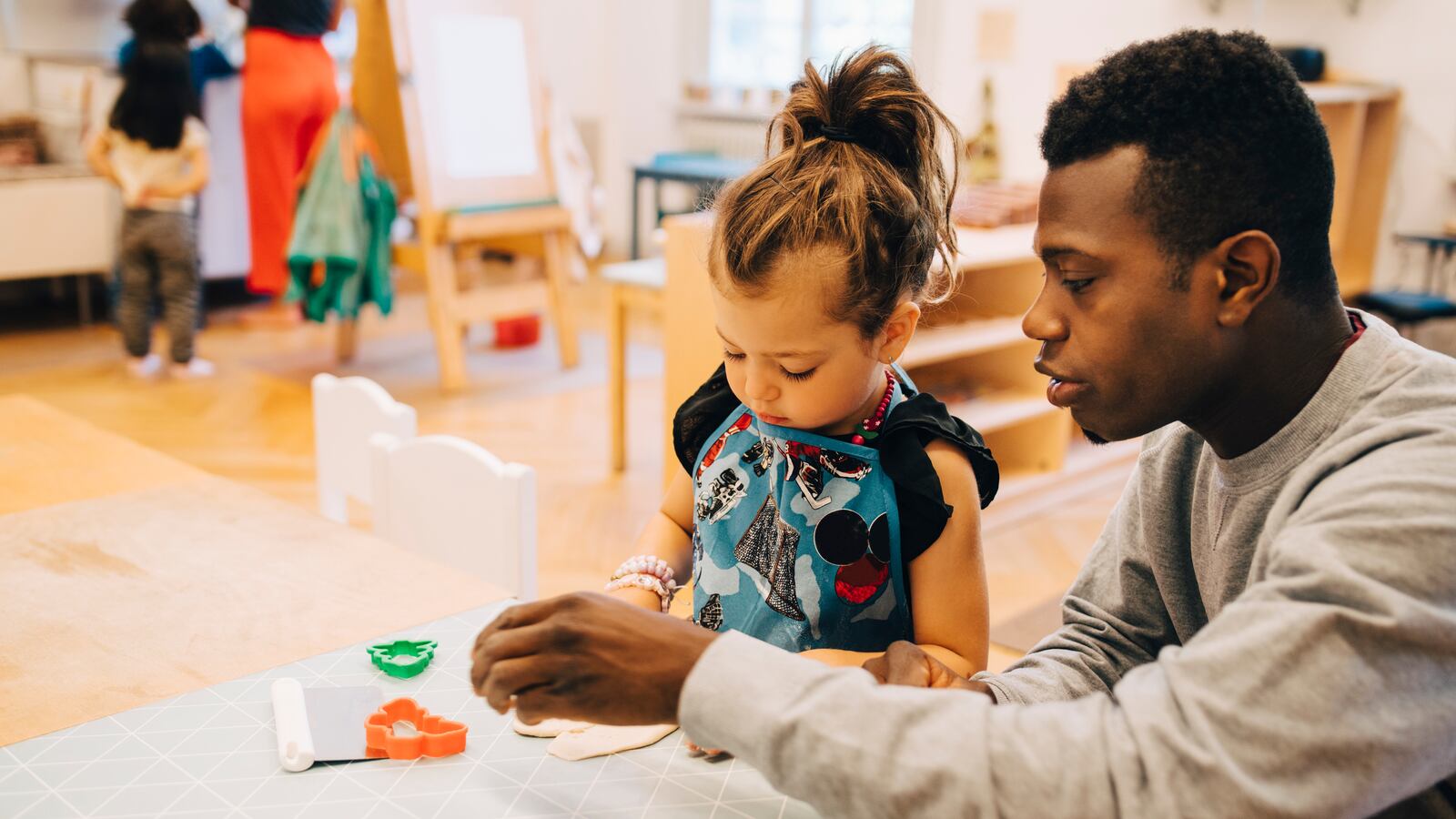 Photo of a male teacher assisting a young girl in a preschool classroom.