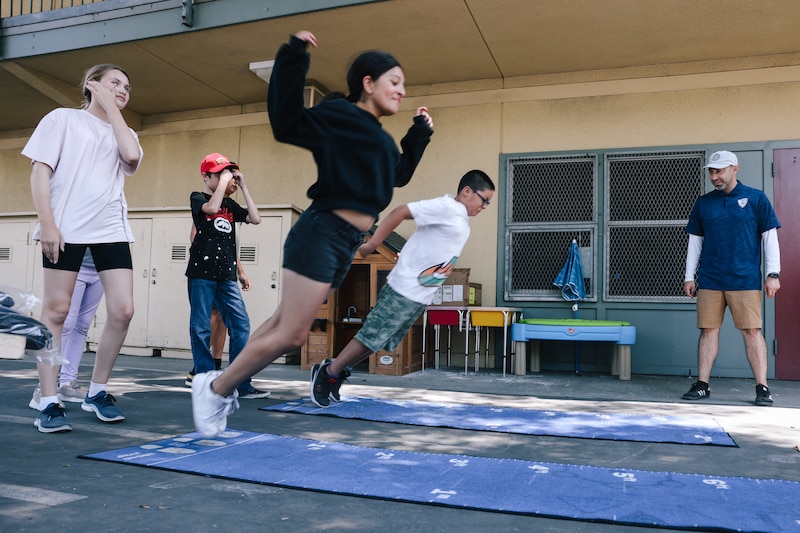 A girl in a black top and shots jumps on a playground while other students surround her.