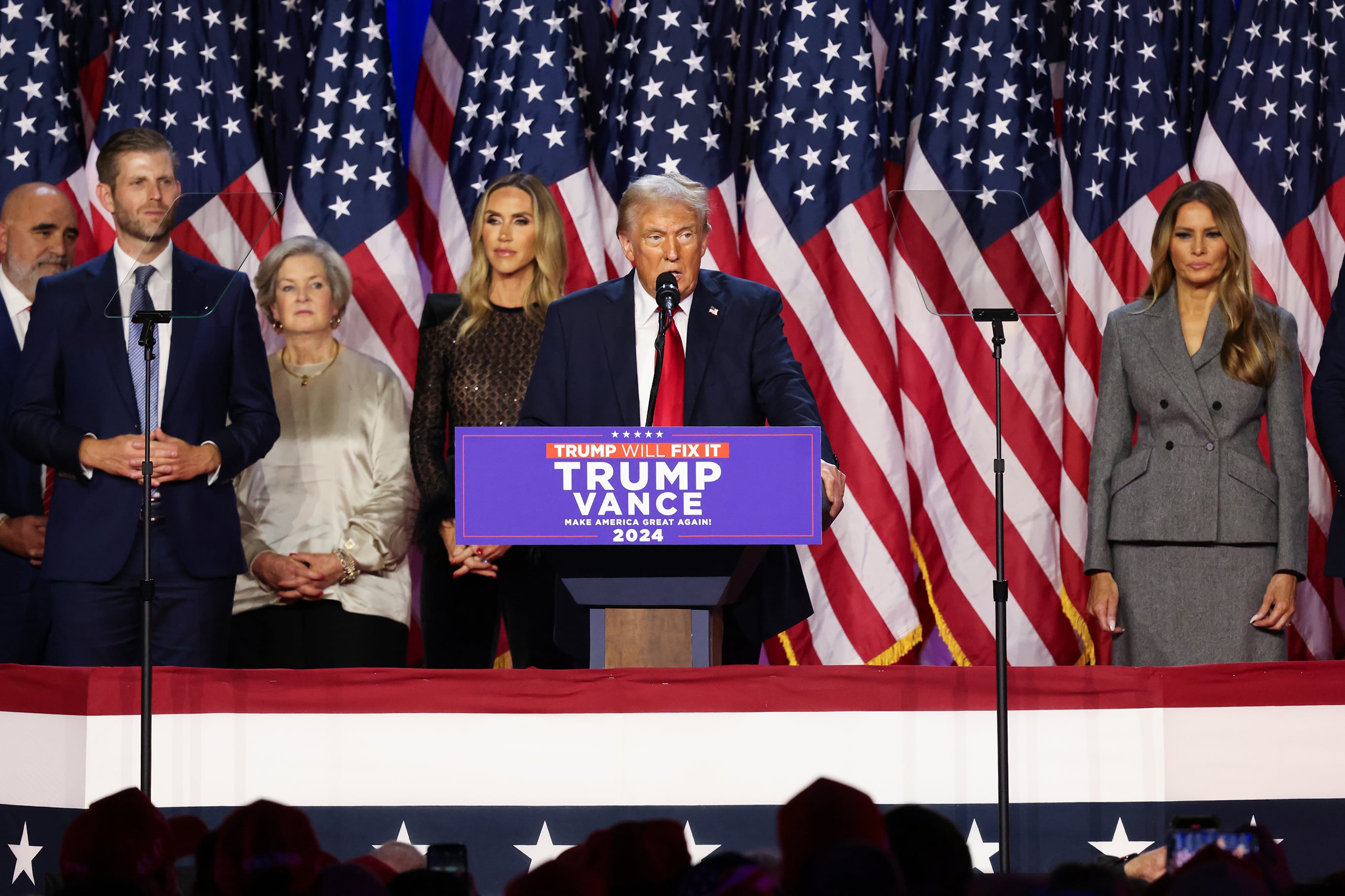 A man in a suit stands behind a podium with a group of people standing behind him and all in front of a row of American flags.